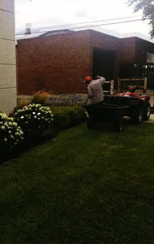 Person mowing a lawn beside a brick building and flower beds.