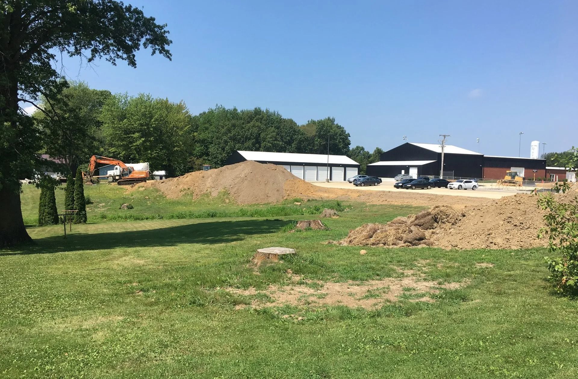 Construction site with piles of dirt, a small excavator, and a building on a sunny day.