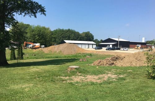 Construction site with piles of dirt, a small excavator, and a building on a sunny day.
