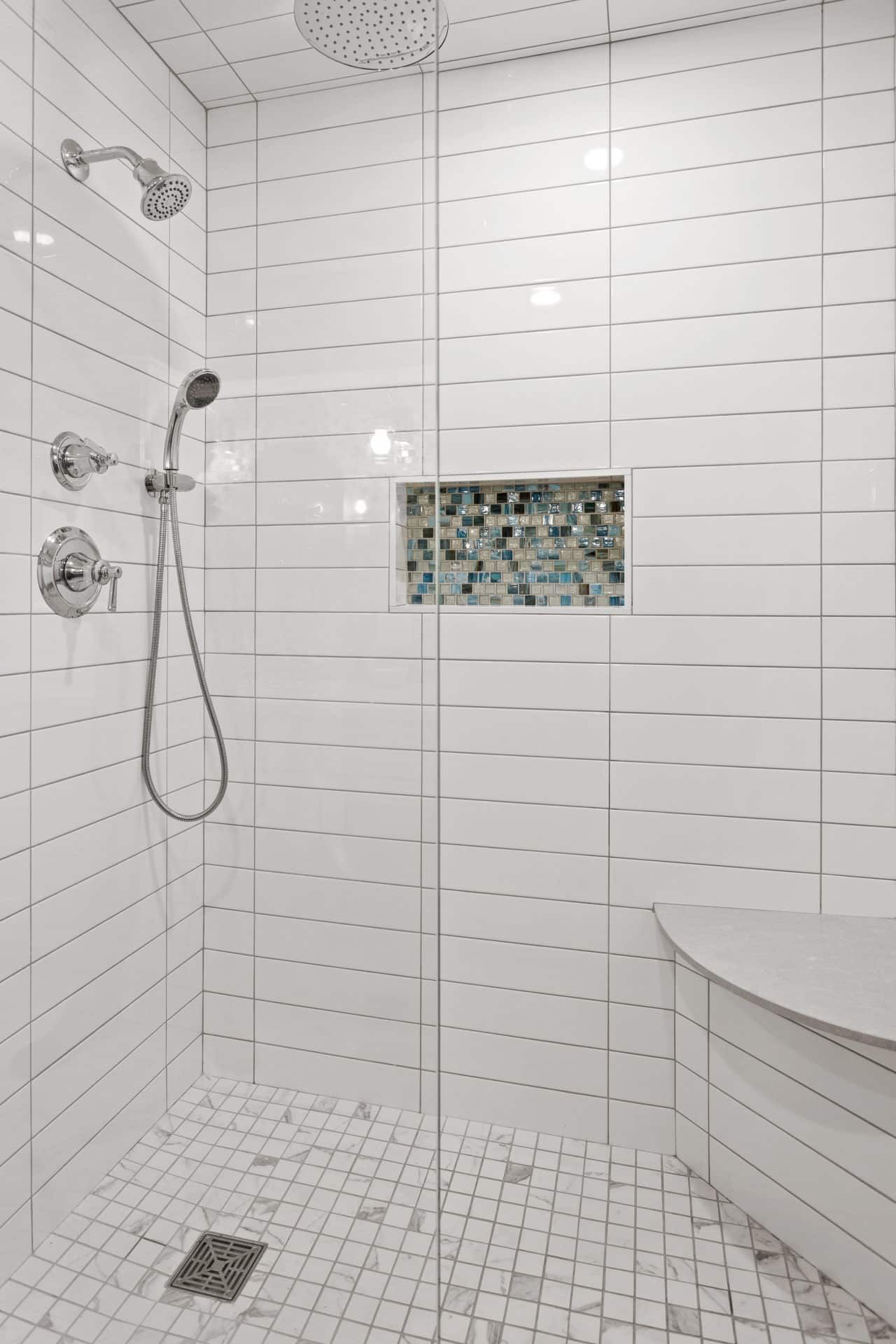 A bathroom with white tiles and a walk in shower with a glass door.