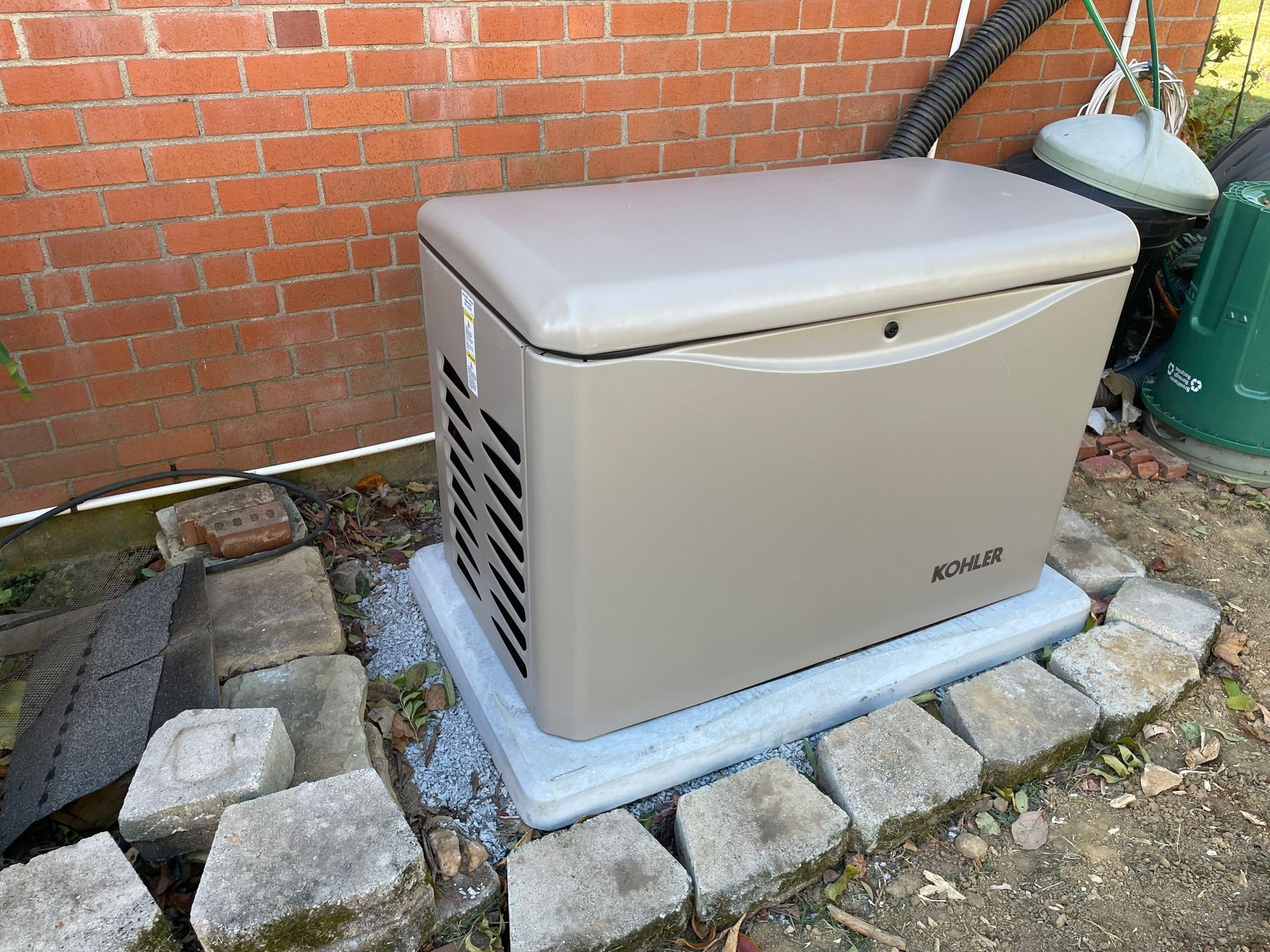 Outdoor beige generator beside a brick wall, sitting on a concrete pad amid stones and grass.