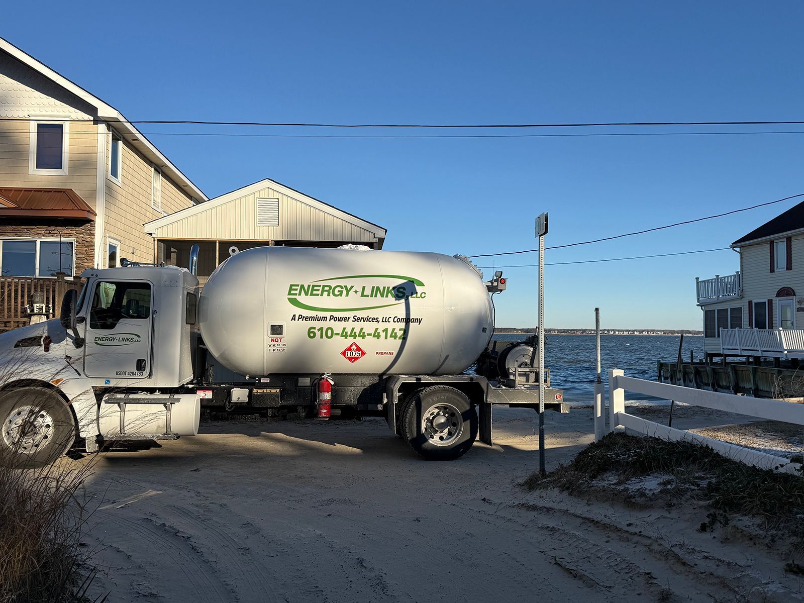 White propane truck on a snowy coastal street beside houses and the ocean, with a lighthouse logo on the tank