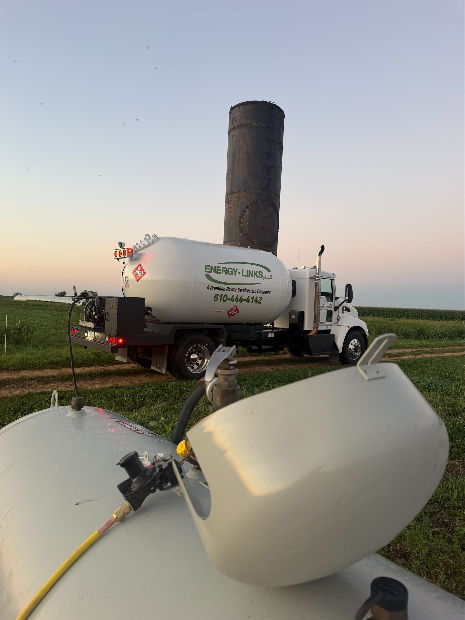 White tanker truck spraying fields at sunset, with a tall cylindrical chimney in the background.