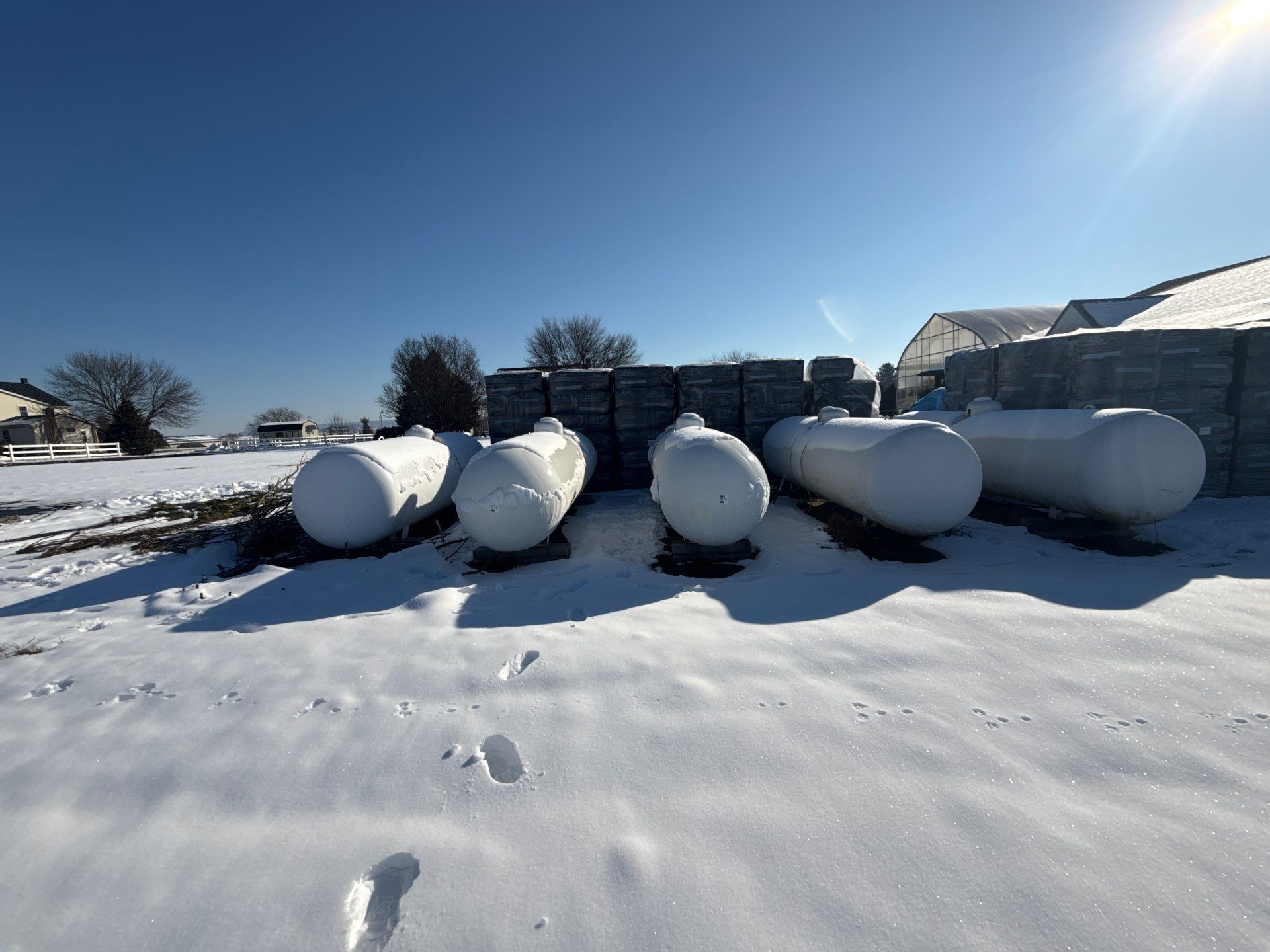 Snow-covered field with several white cylindrical hay bales near farm buildings under a bright blue sky