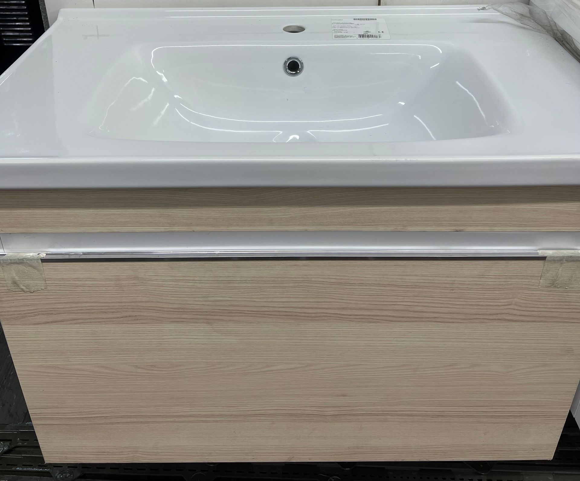 A modern wall-mounted bathroom vanity with a white ceramic sink and light wood-grain cabinet featuring a metal handle.
