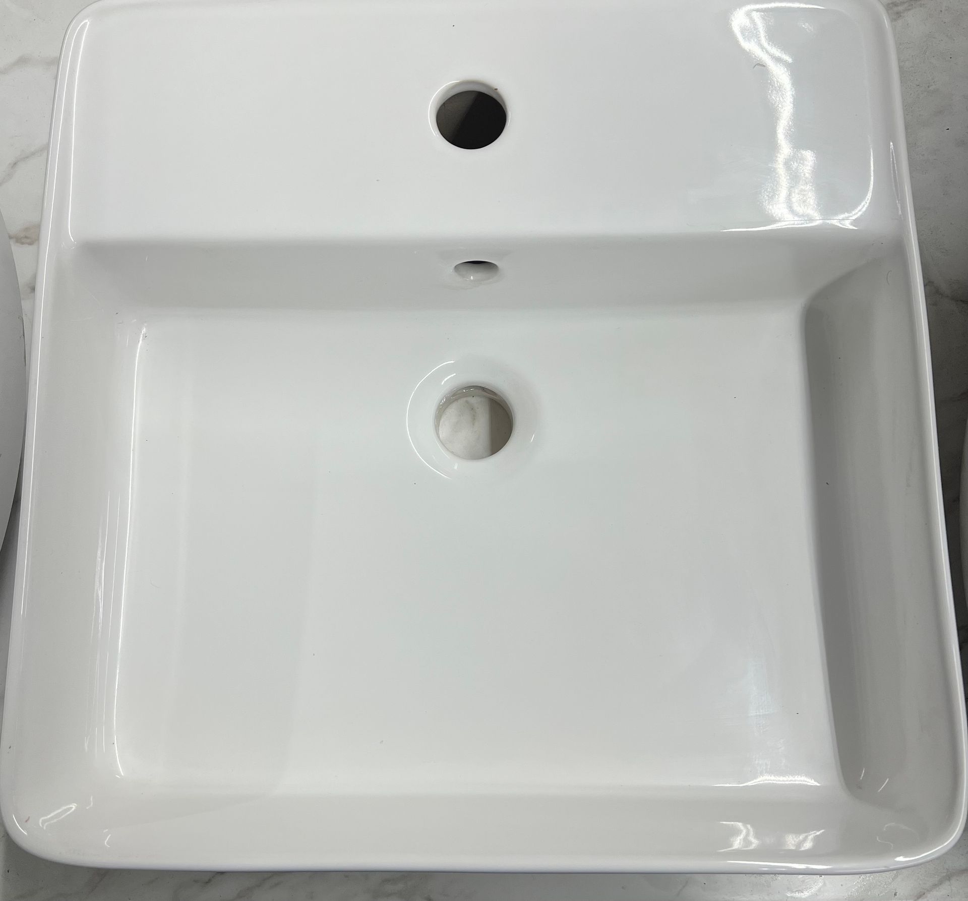 A square, white ceramic bathroom sink with a centered faucet hole, an overflow, and a central drain opening.