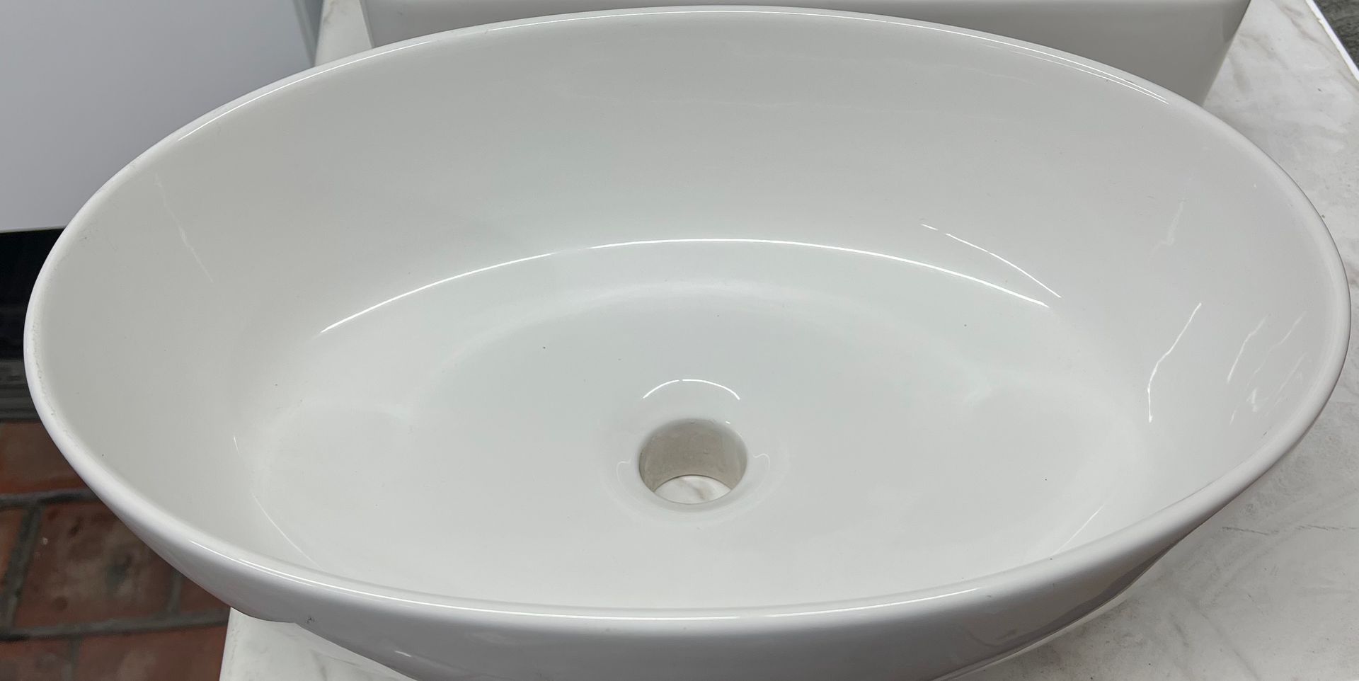 An oval, white ceramic vessel bathroom sink with a central drain hole, set against a blurred background.
