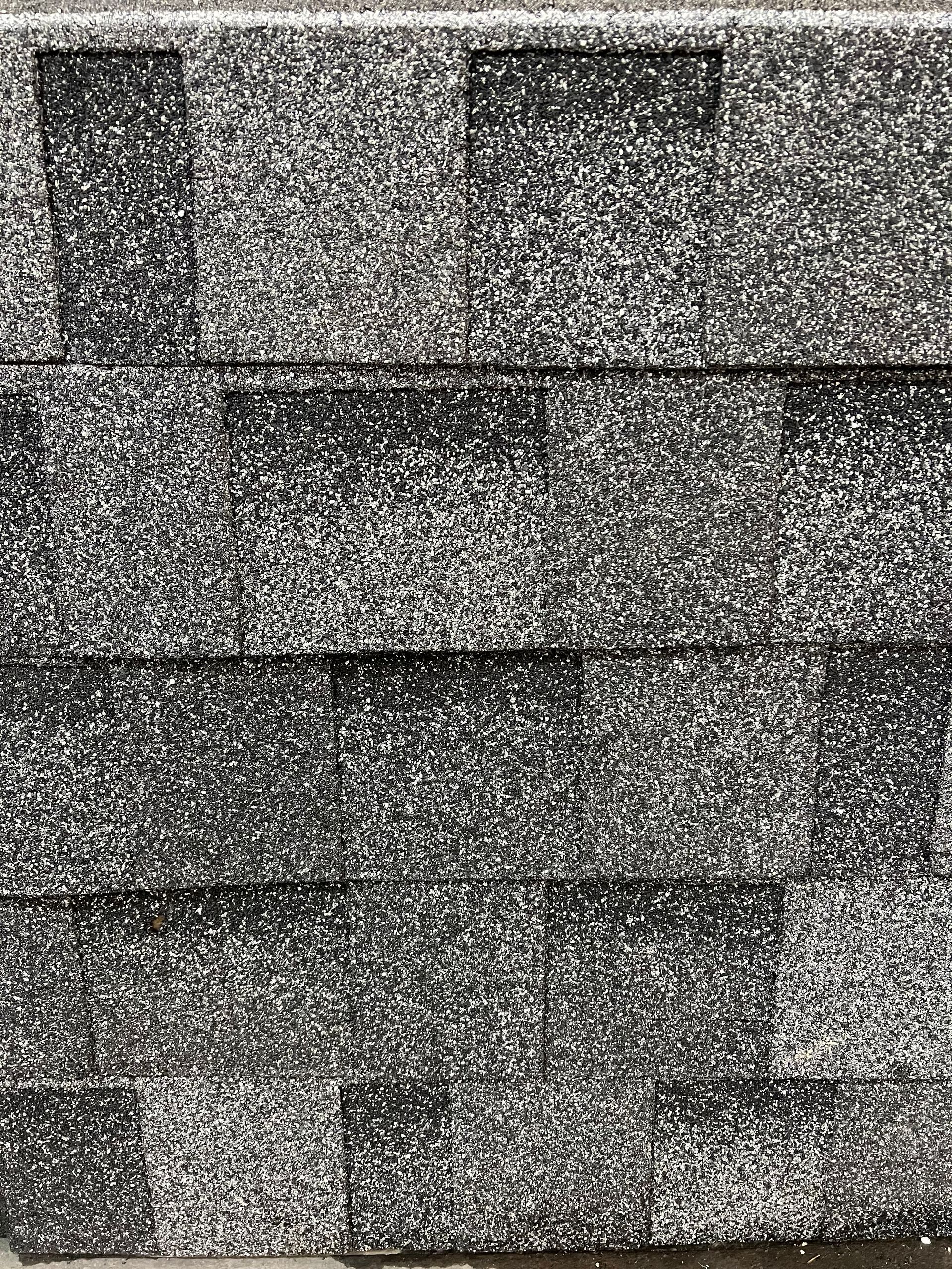 A close-up of dark grey, black, and white asphalt roof shingles with a multi-layered, textured surface.