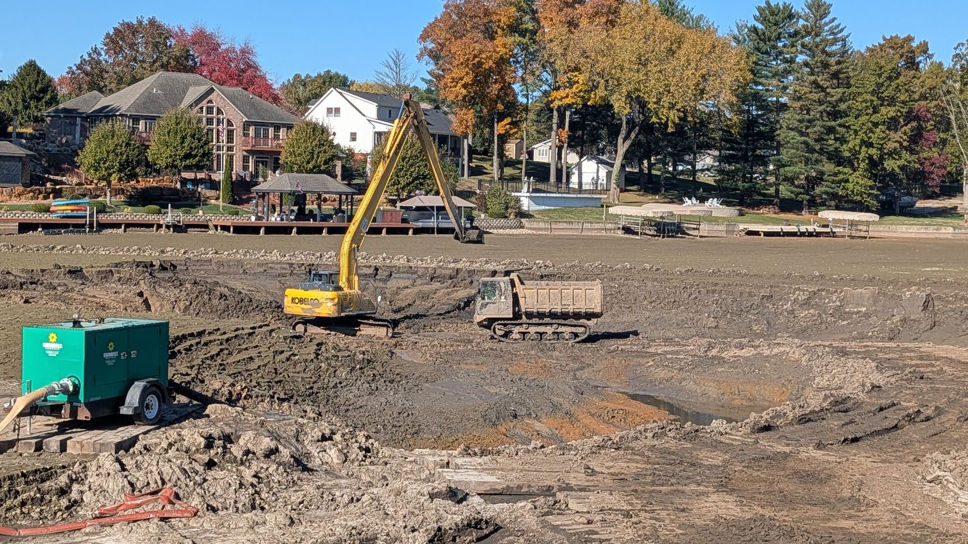 Construction site: Excavator and dump truck in a drained lakebed, residential houses in the background.