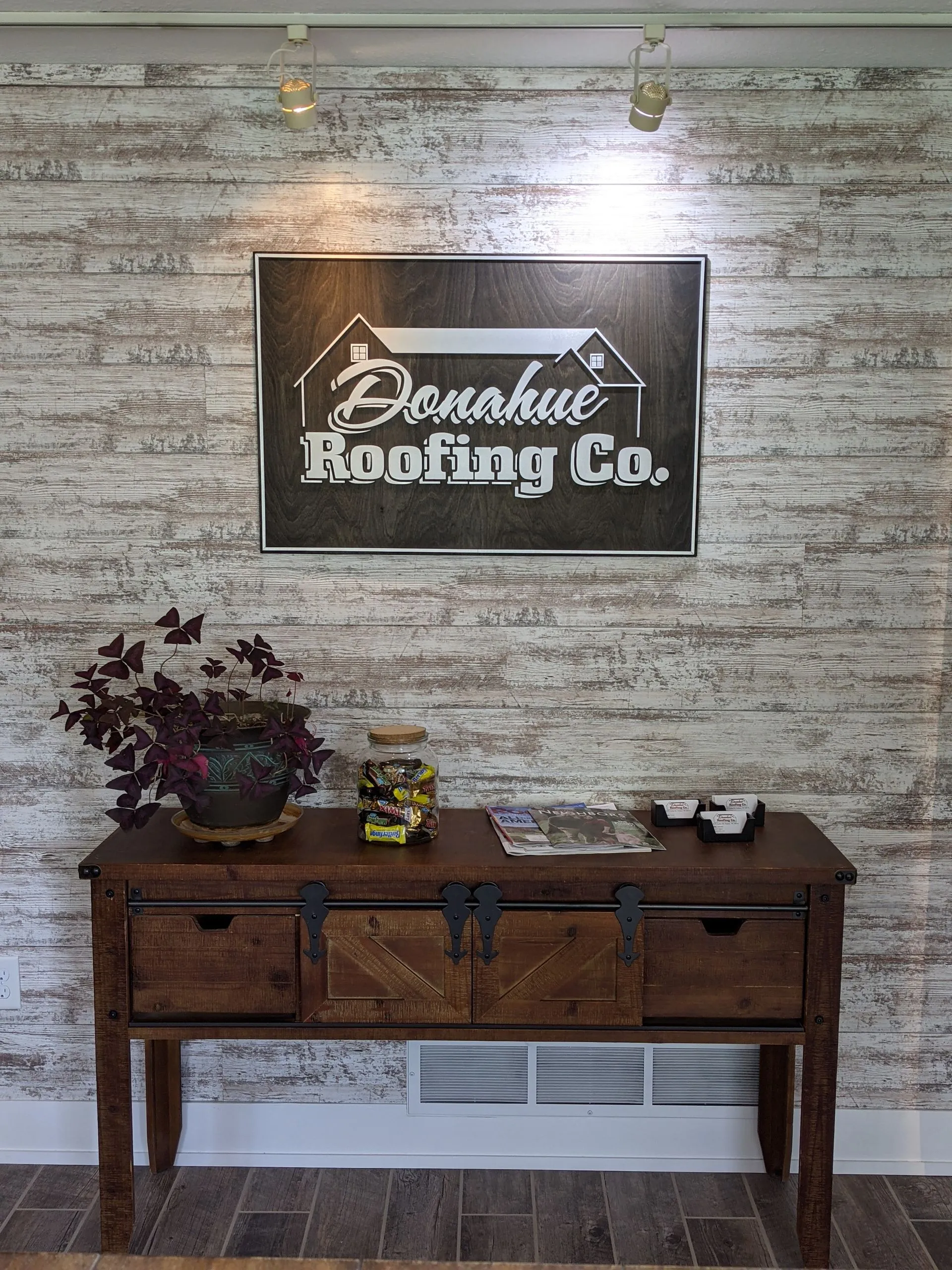 A wooden table with a plant and company sign on a brick-like wall. The sign reads