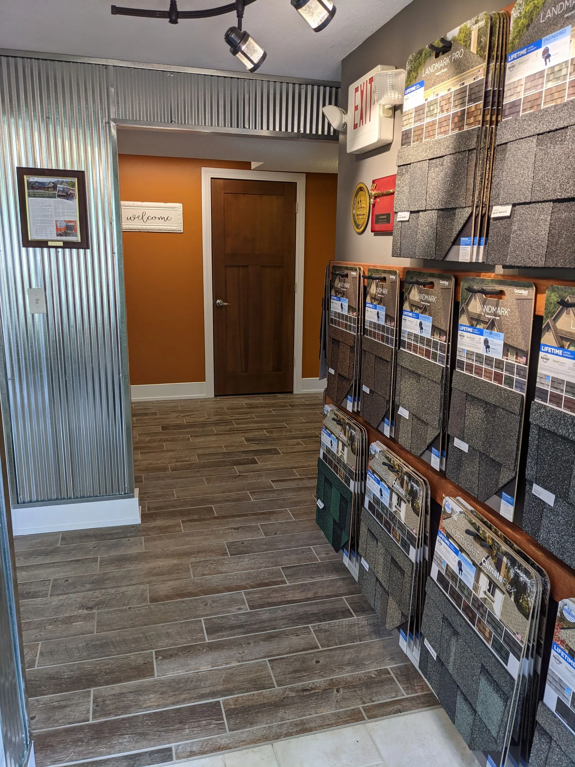 Interior of a showroom displays various roofing samples and tile flooring. A doorway and hallway are visible.