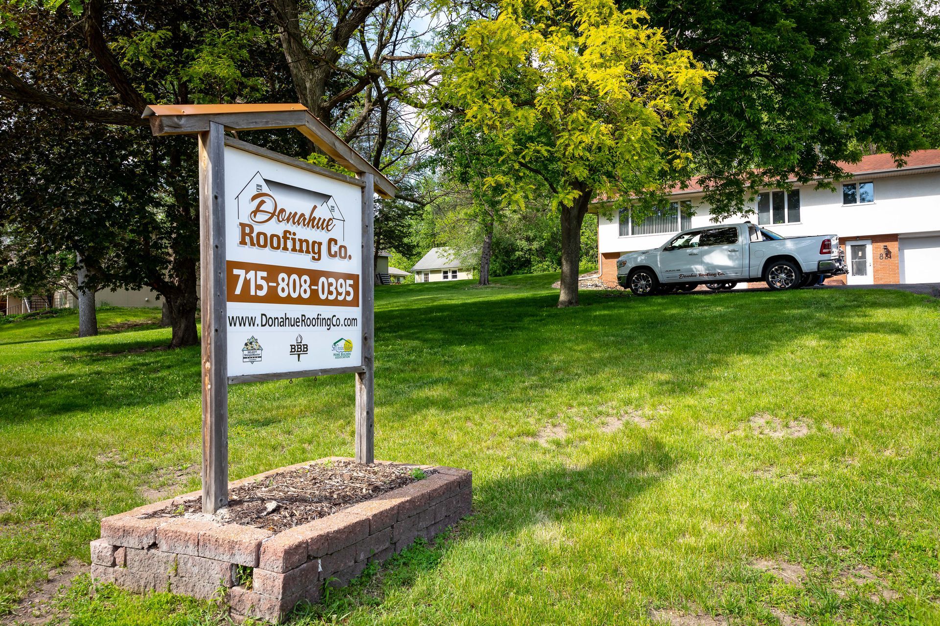 A sign for a roofing company is sitting in the grass in front of a house.