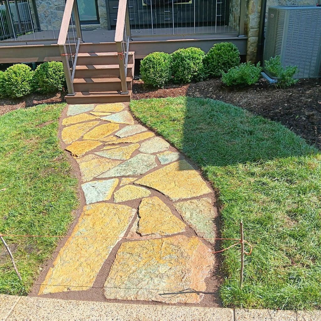 Stone walkway leading to a house entrance, bordered by grass and shrubs, with steps and a porch in view