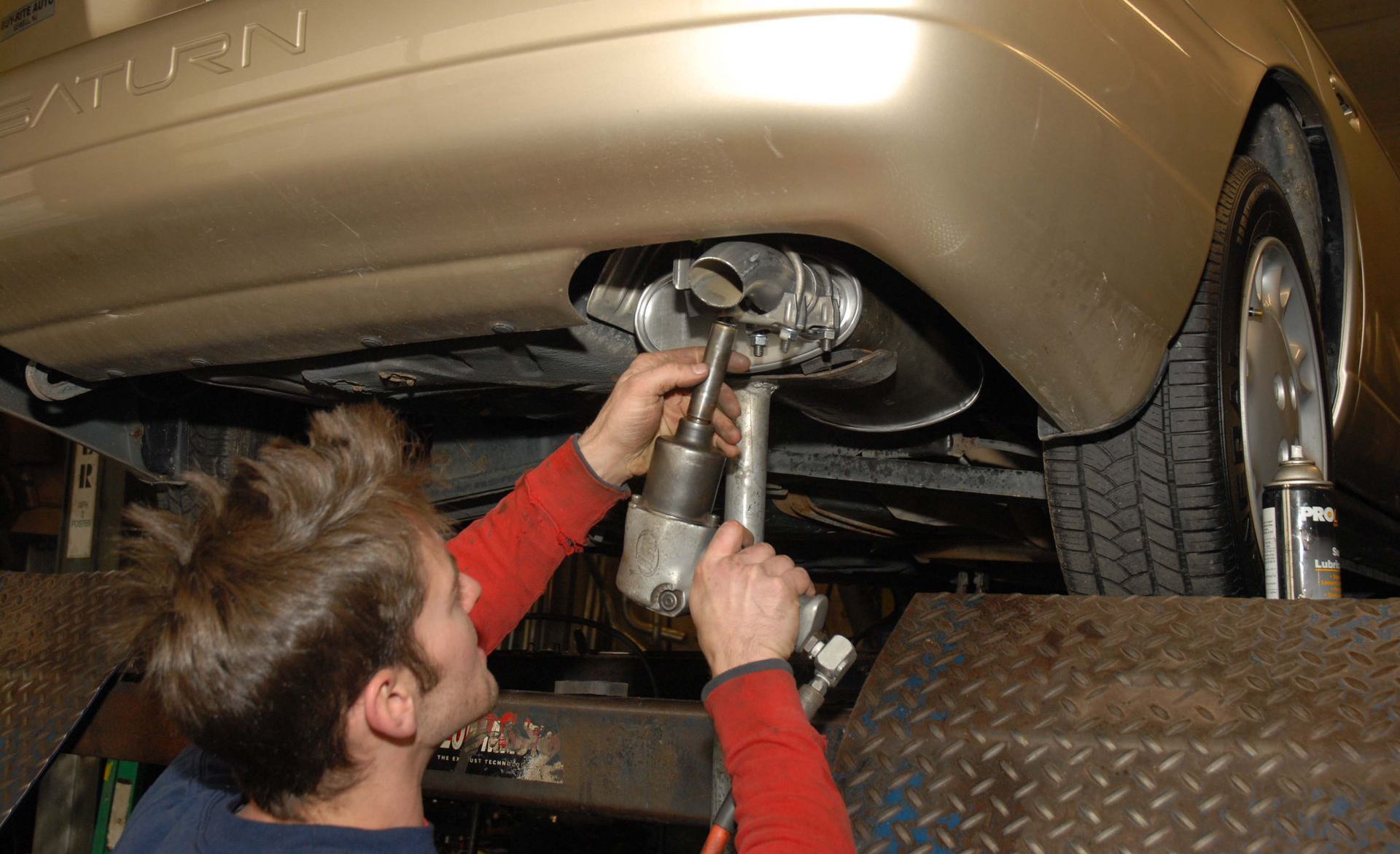 Mechanic working under a gold car on a lift, using a tool near the exhaust.