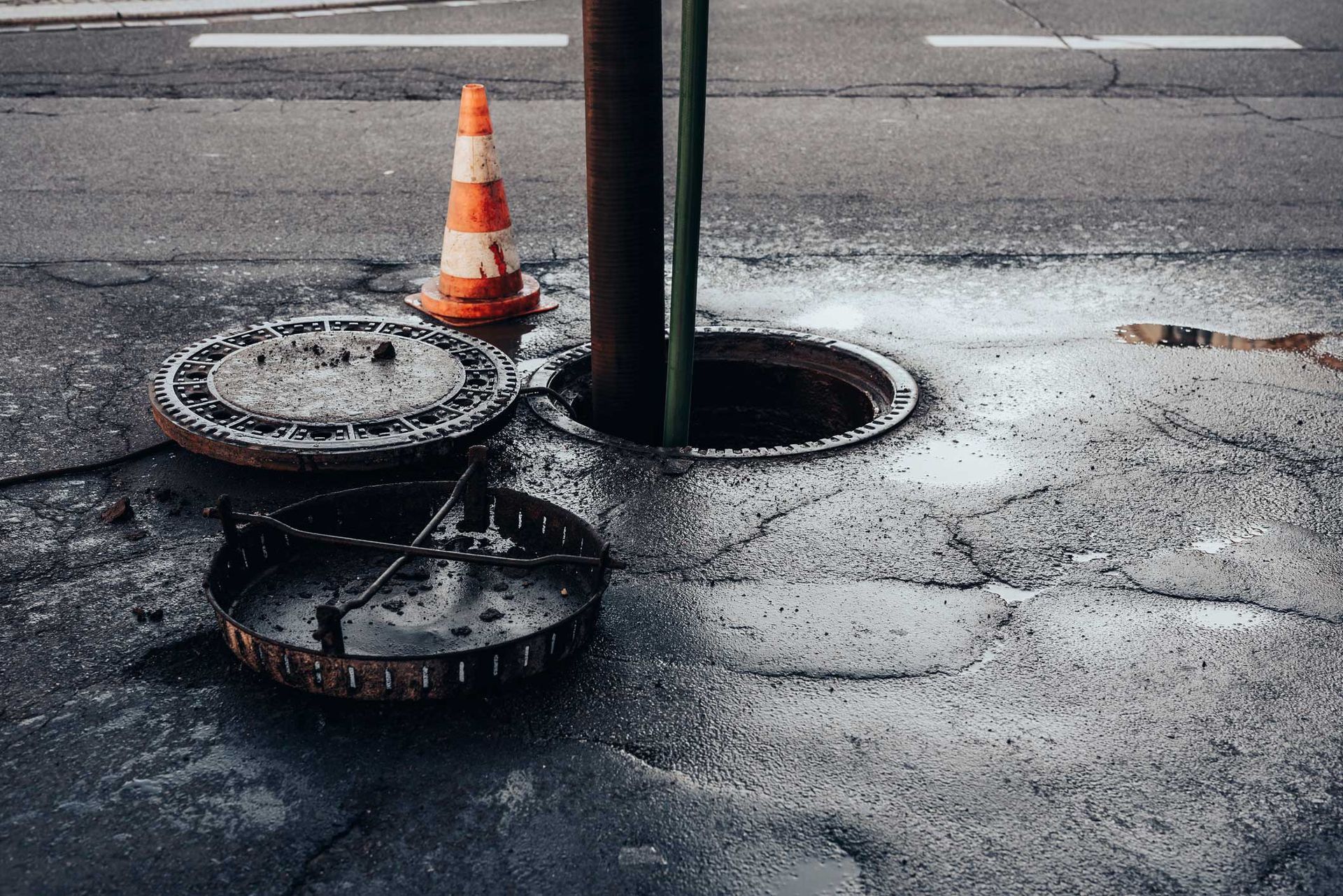 Open sewer on street with open manhole, cone, metal cover, and pole.