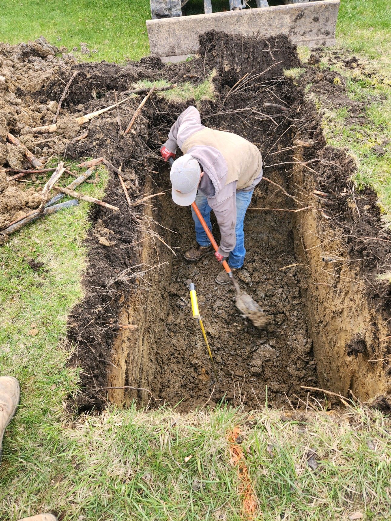 Person digging in a rectangular hole in the ground with a shovel; surrounding ground is grassy and muddy.