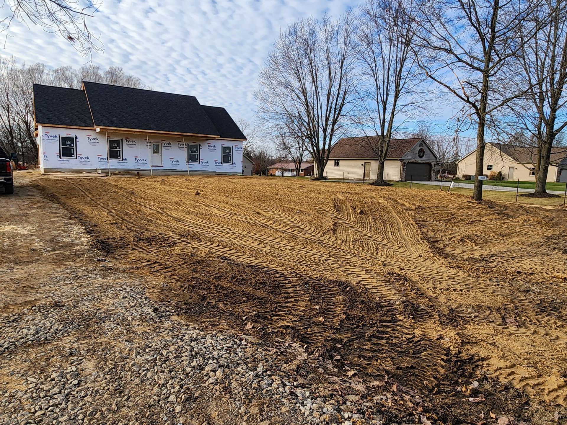 New house under construction with brown dirt yard. White wrap on the house. Blue sky with bare trees.