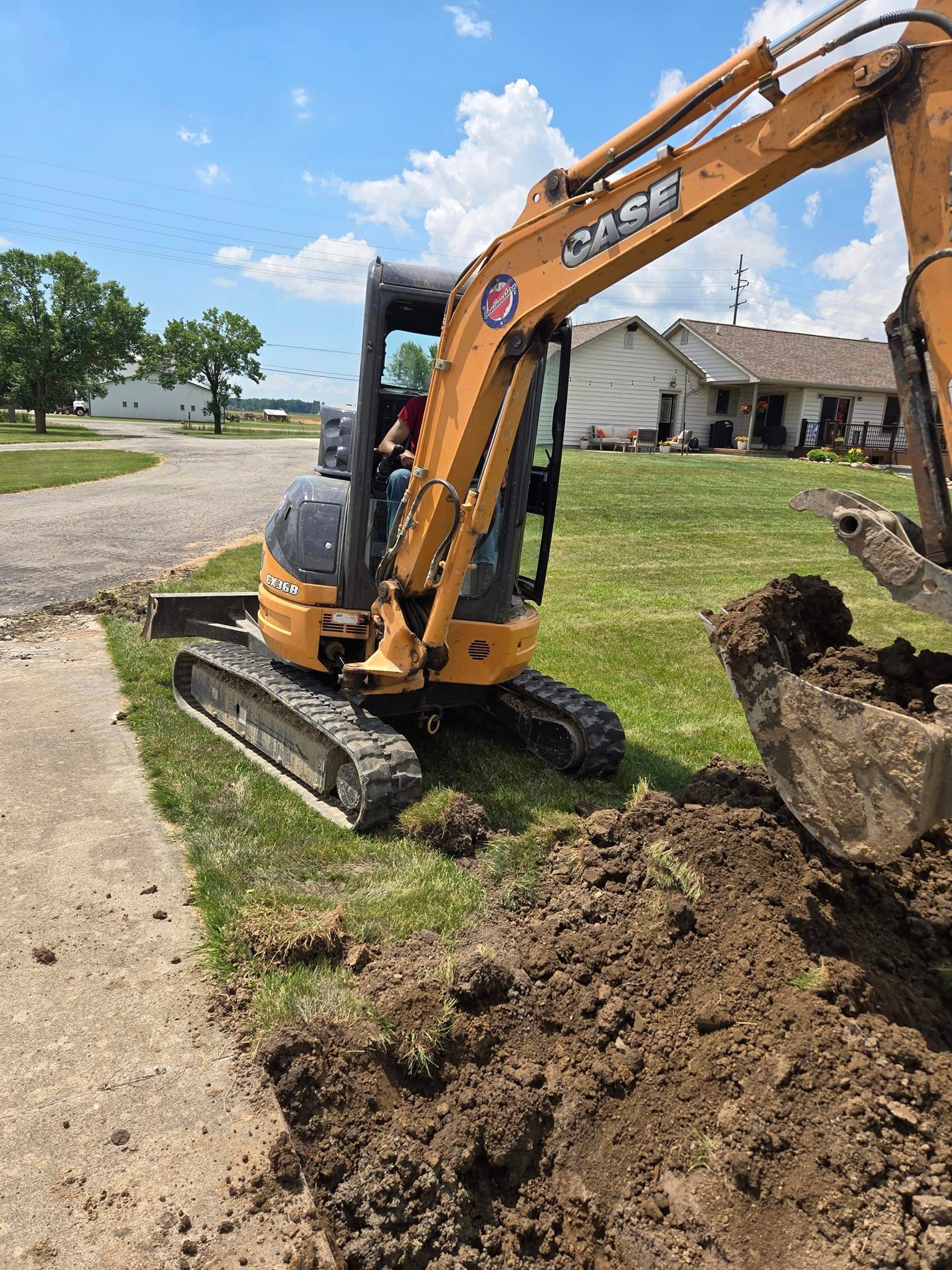 Small orange Case excavator digging a trench in a grassy yard near a sidewalk and houses.