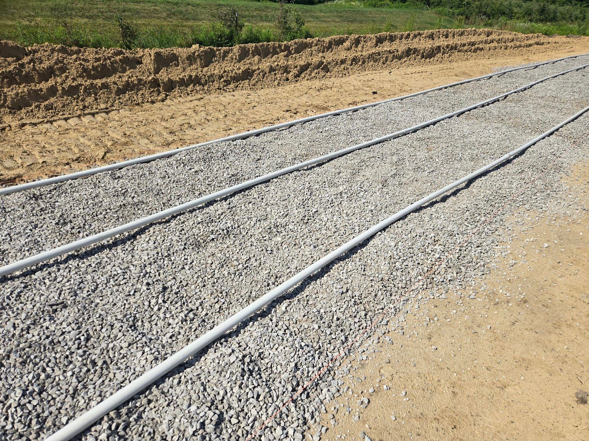 Three gray pipes laid on gravel path, adjacent to a dirt area, with green vegetation in the background.