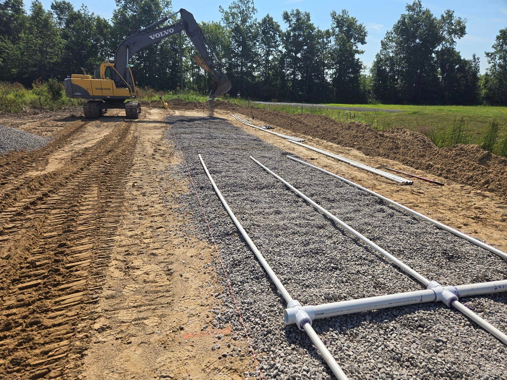 Construction of a septic system: excavator working on gravel-filled trench with white pipes for drainage.