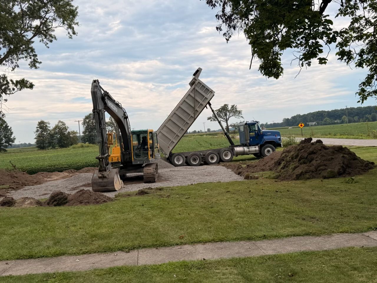 Excavator loading soil into a dump truck at a construction site near a grassy field and road