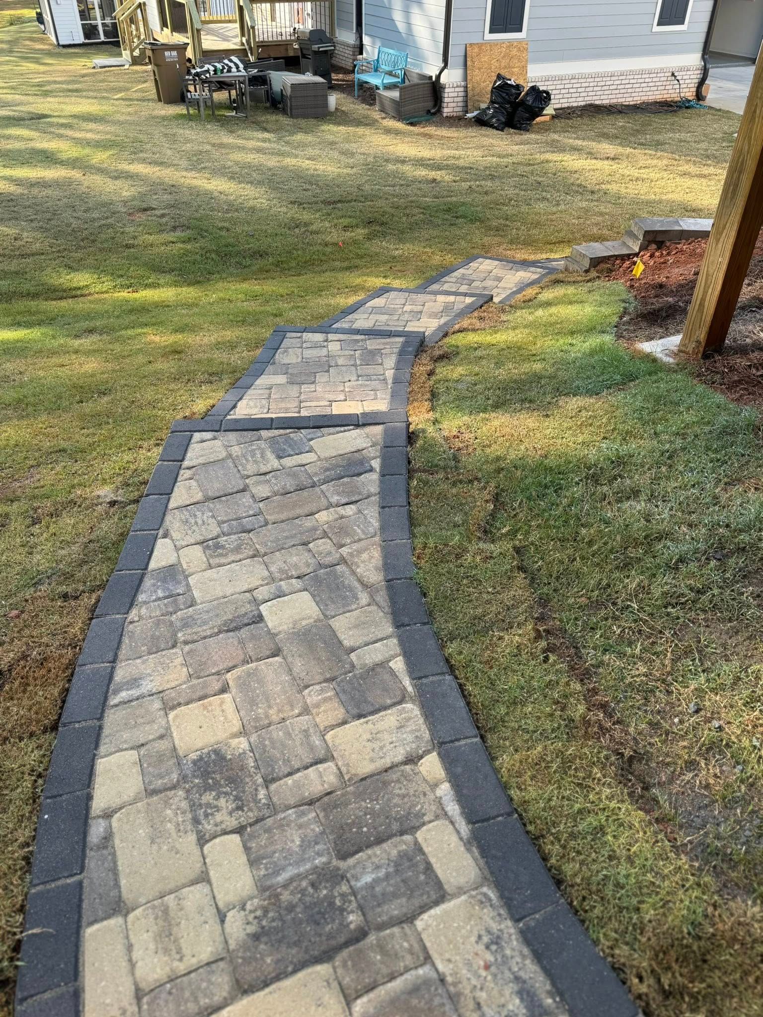 Brick path with black border winds through a grassy yard, leading towards a house.
