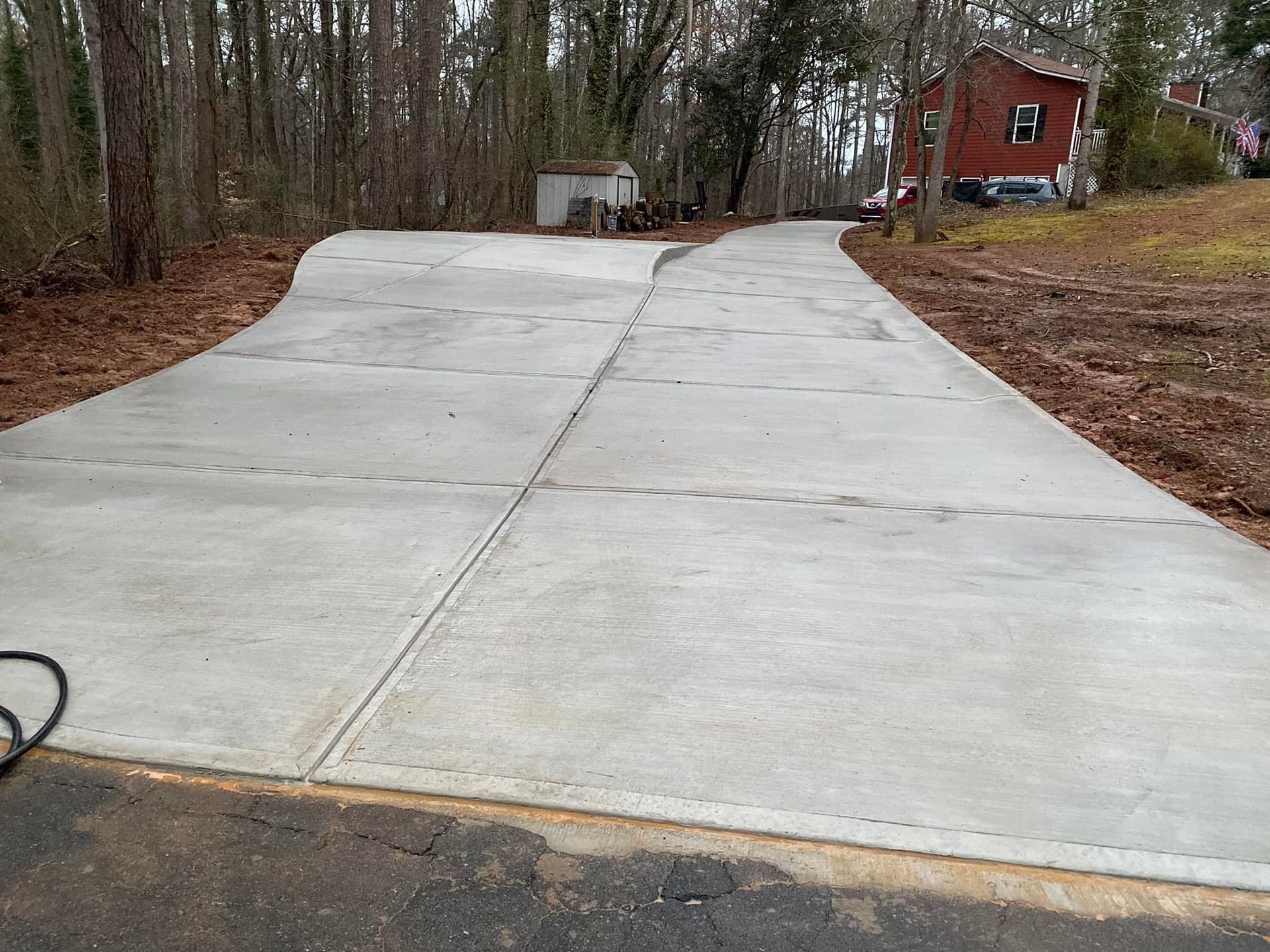 Newly poured concrete driveway curves up a wooded hill toward a red house; small shed visible.