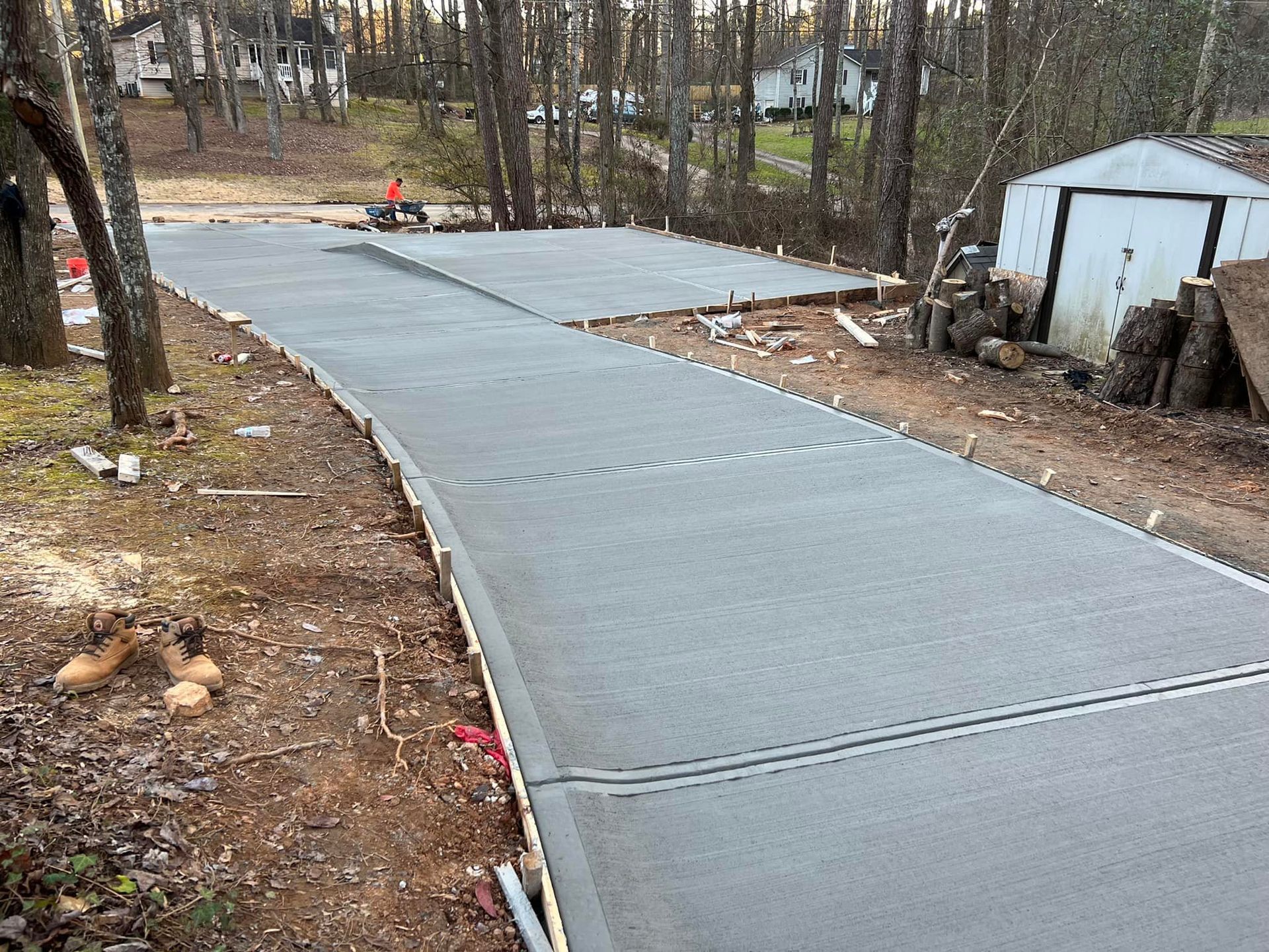 Newly poured concrete driveway and pad, surrounded by dirt and trees, with a small shed in the background.