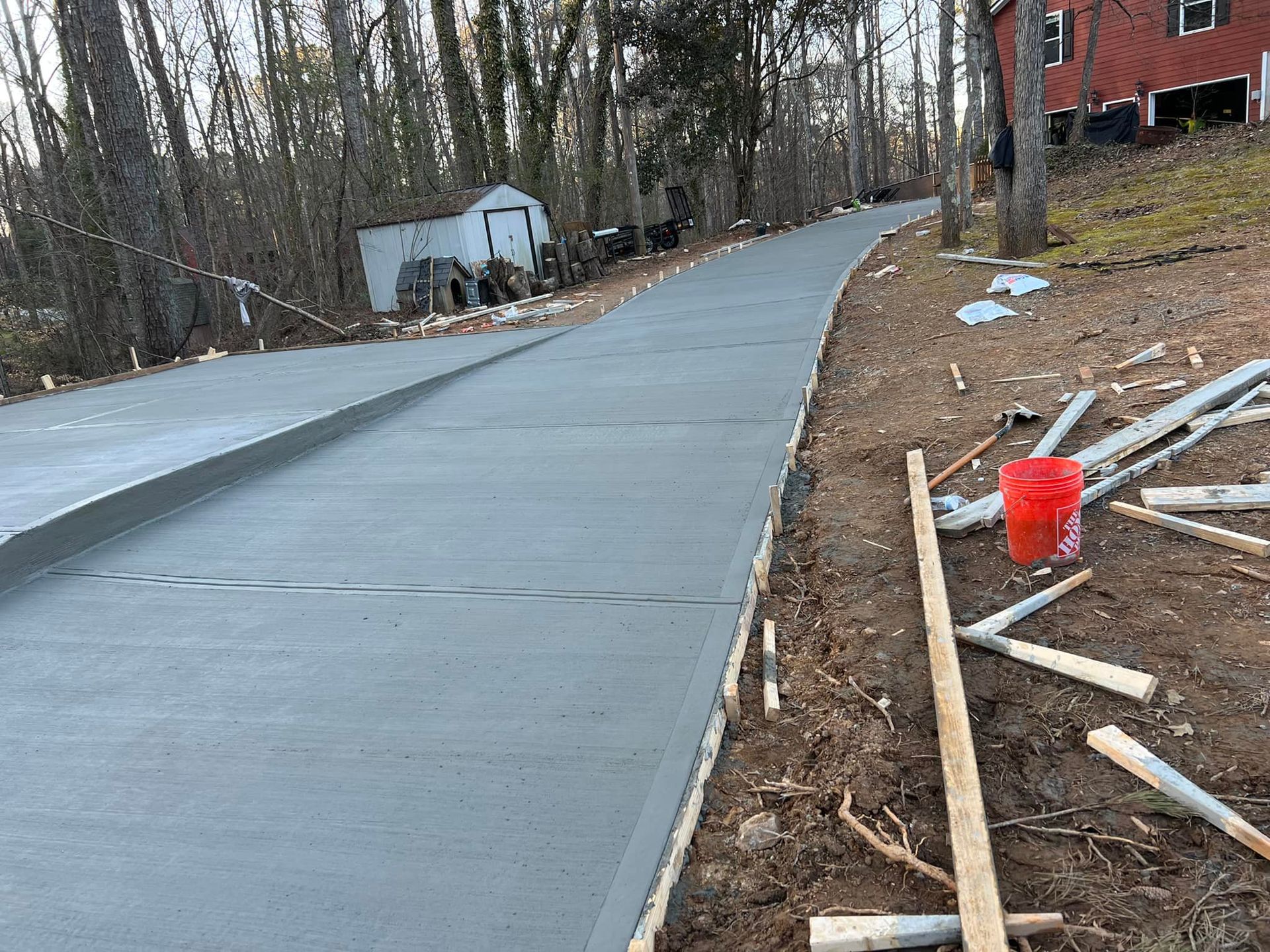 Newly poured concrete driveway extending uphill, next to trees and a red bucket.