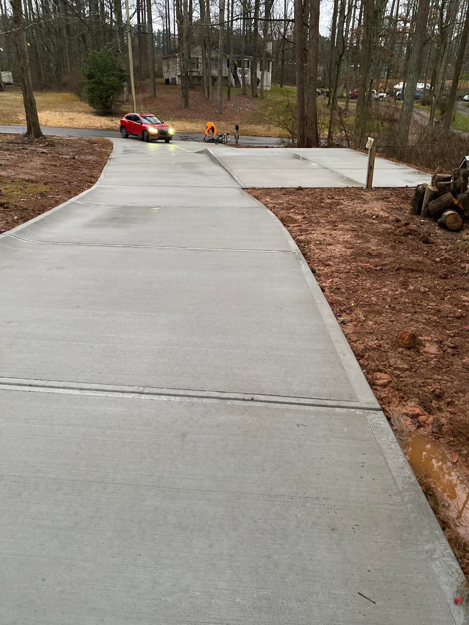 Red car driving on a concrete driveway, next to dirt and trees.