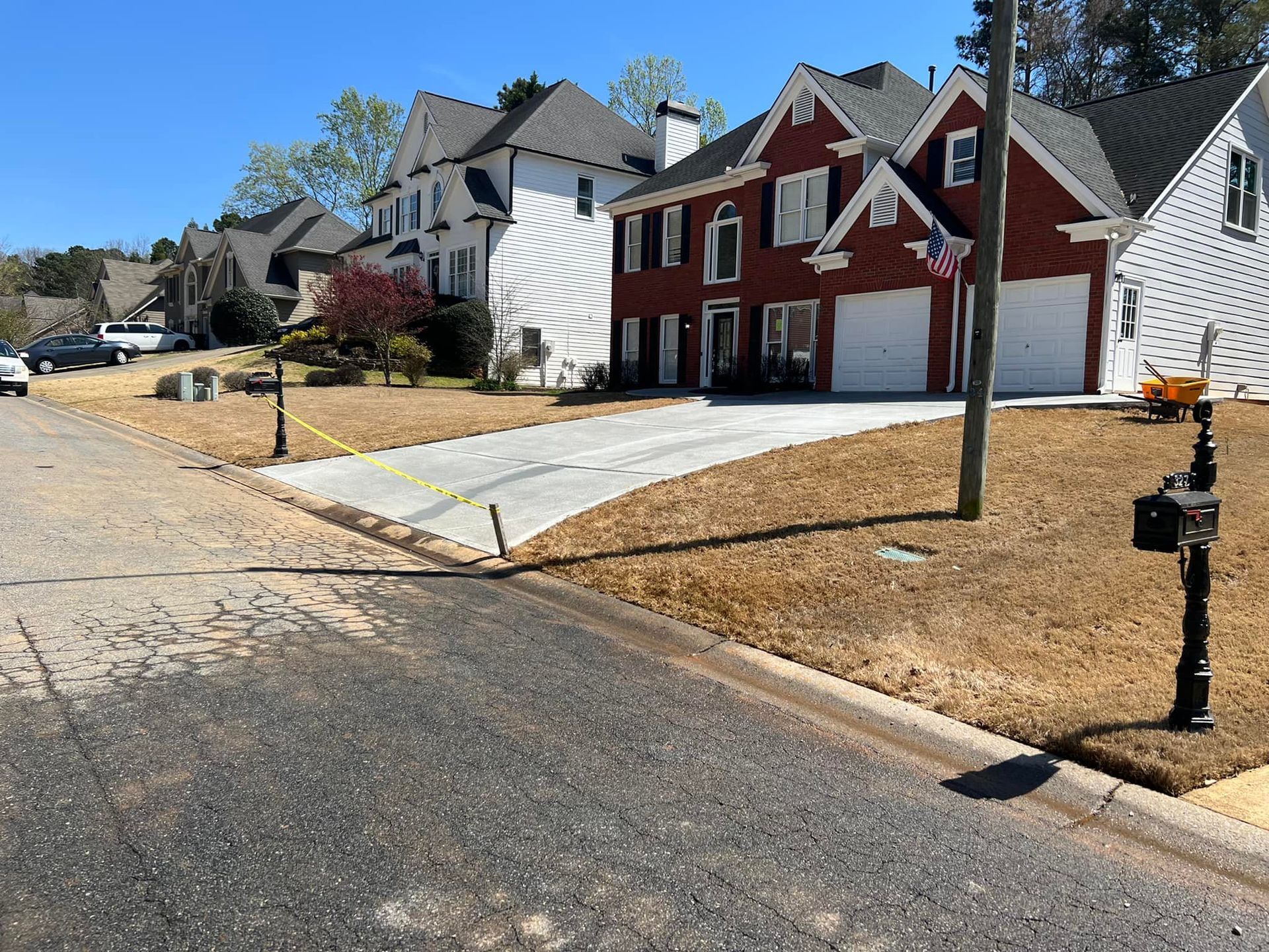 Houses line a street with brown lawns. Red brick house with white garage doors. Blue sky.