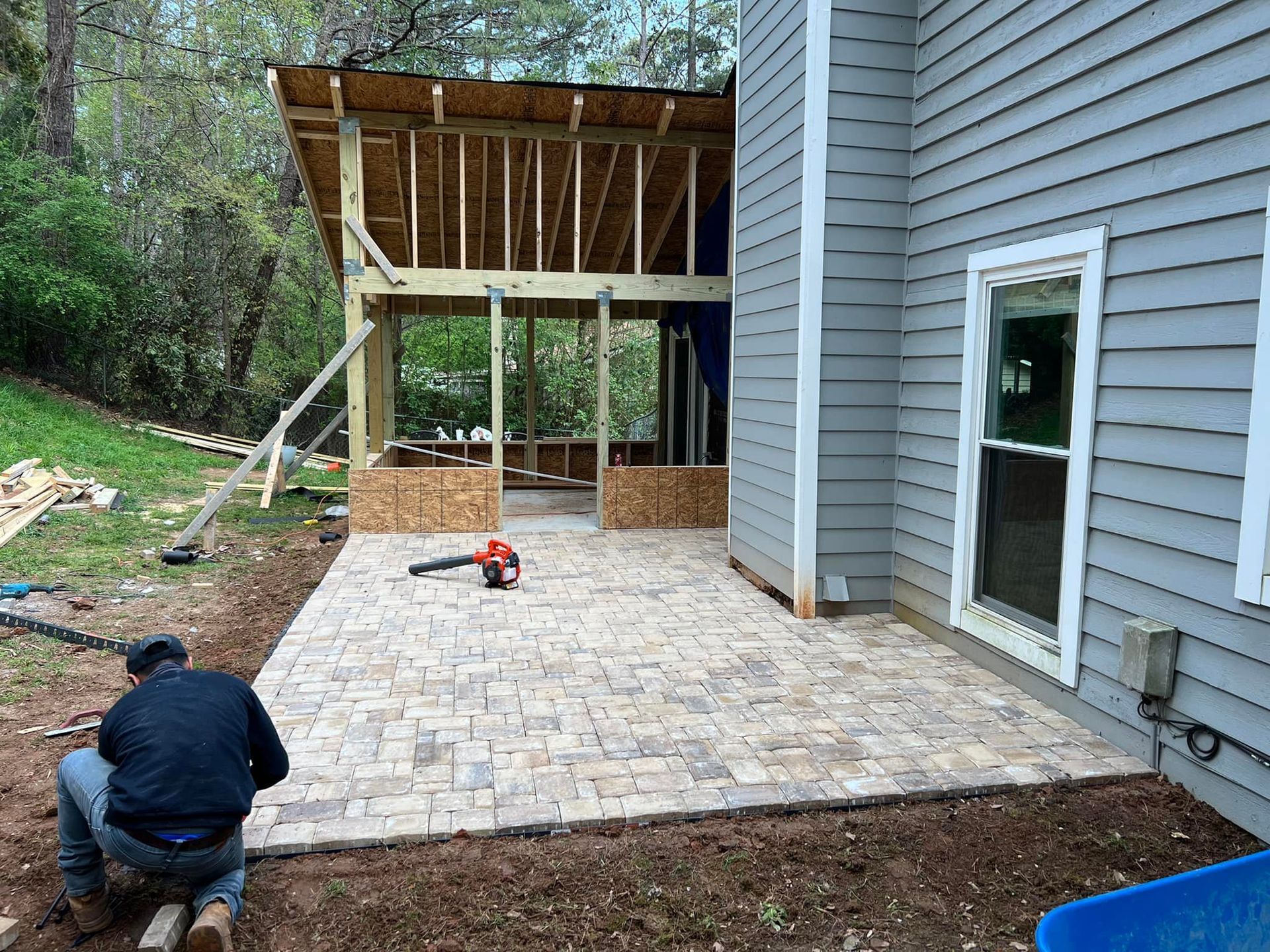 Construction of a patio and screened porch addition to a house. Man laying bricks, unfinished wooden structure.