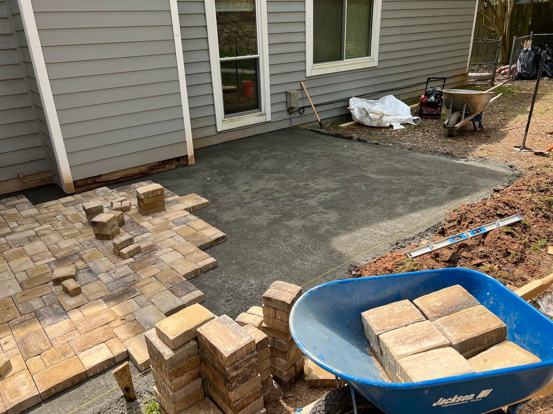 Construction of a patio with pavers; a wheelbarrow, bricks, and a house in the background.