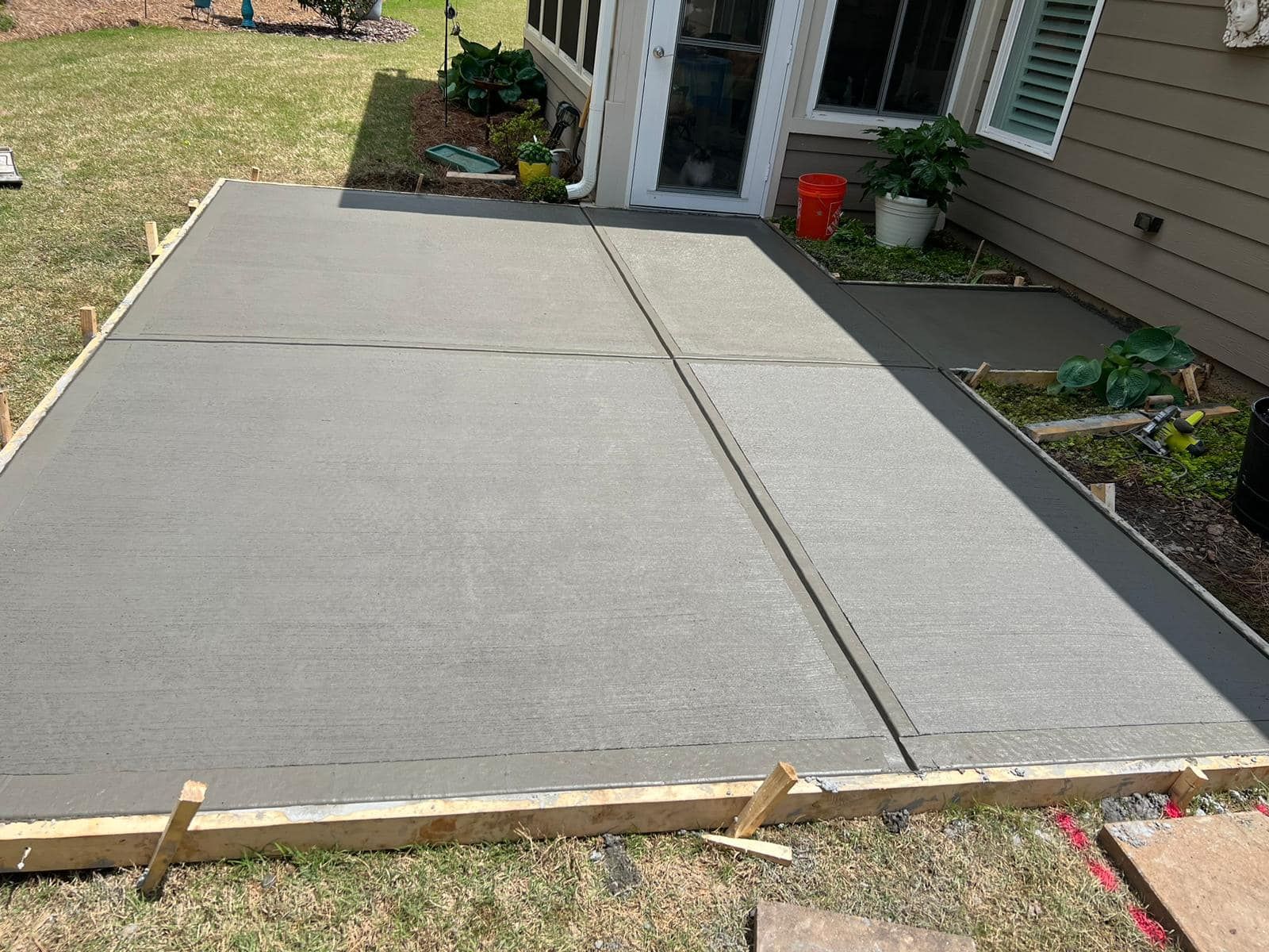 Freshly poured gray concrete patio with wooden borders, next to a house.