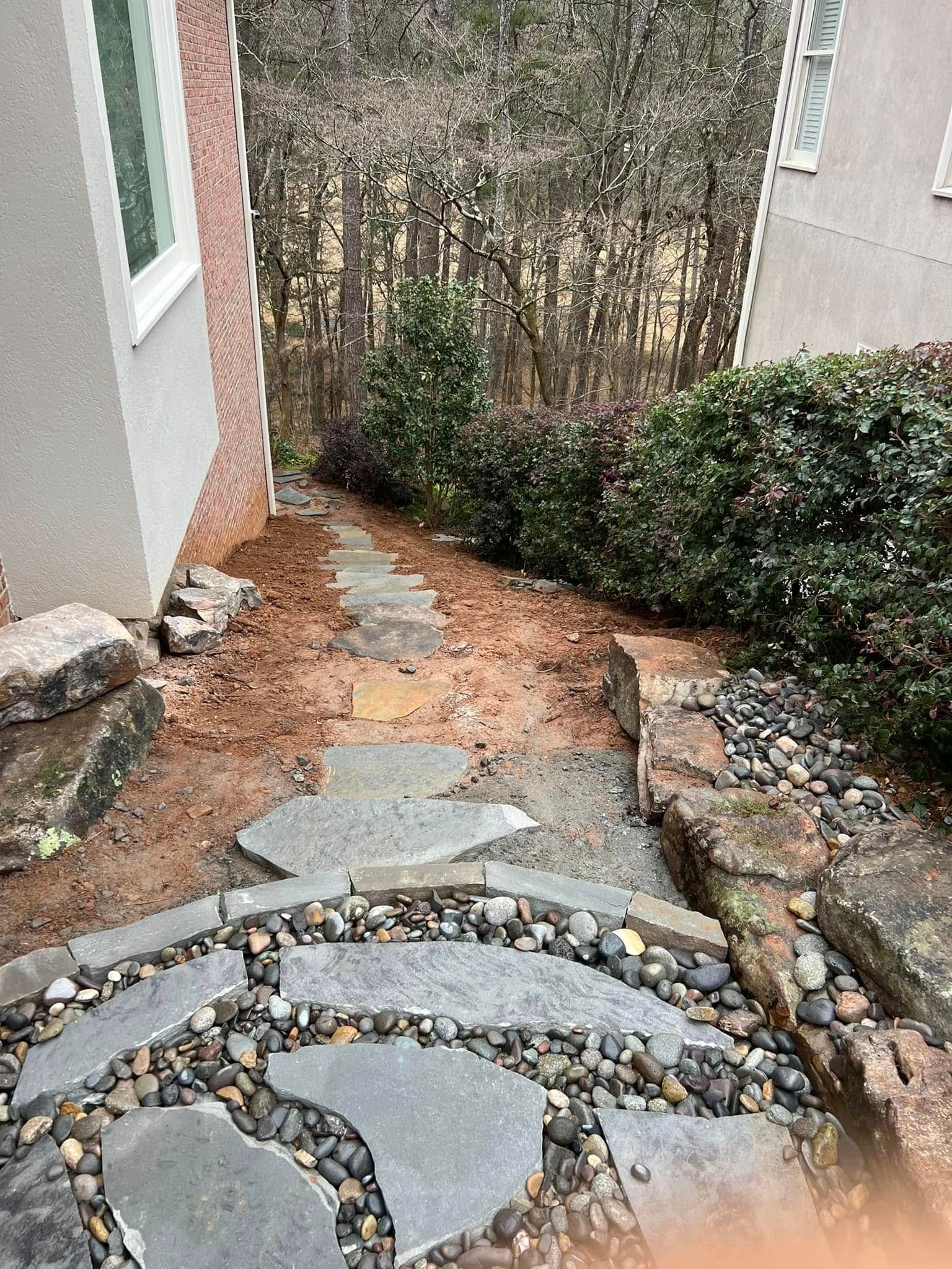 Stone pathway leads through a yard, bordered by rocks, shrubs, and two buildings.
