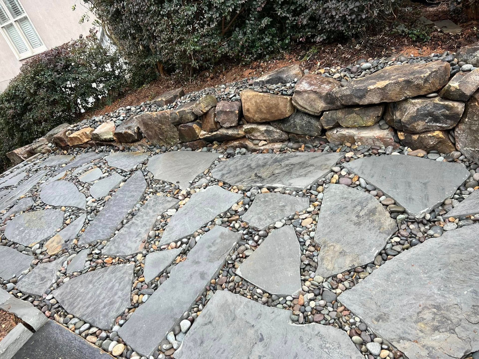 Stone pathway with irregular-shaped pavers and gravel between, bordered by a stone wall and foliage.