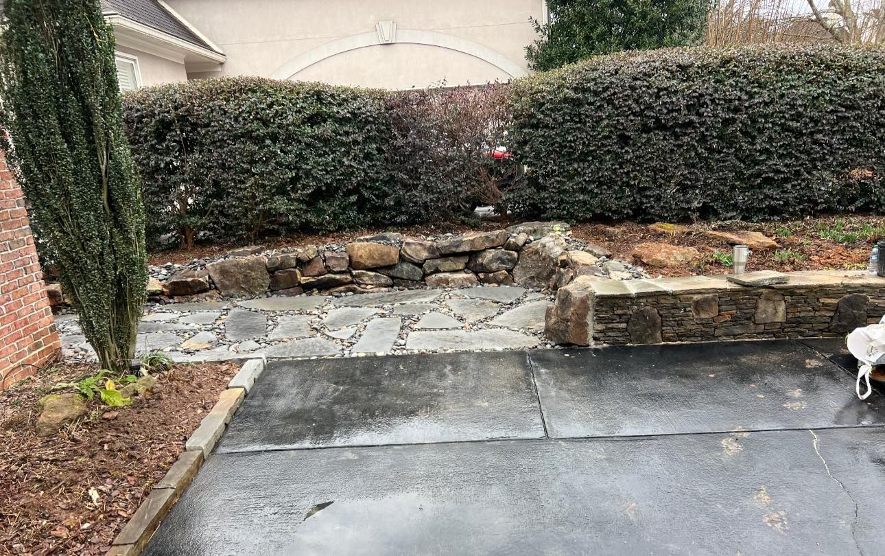 Stone pathway leading to a garden bed with a trimmed green hedge, bordered by a stone wall.