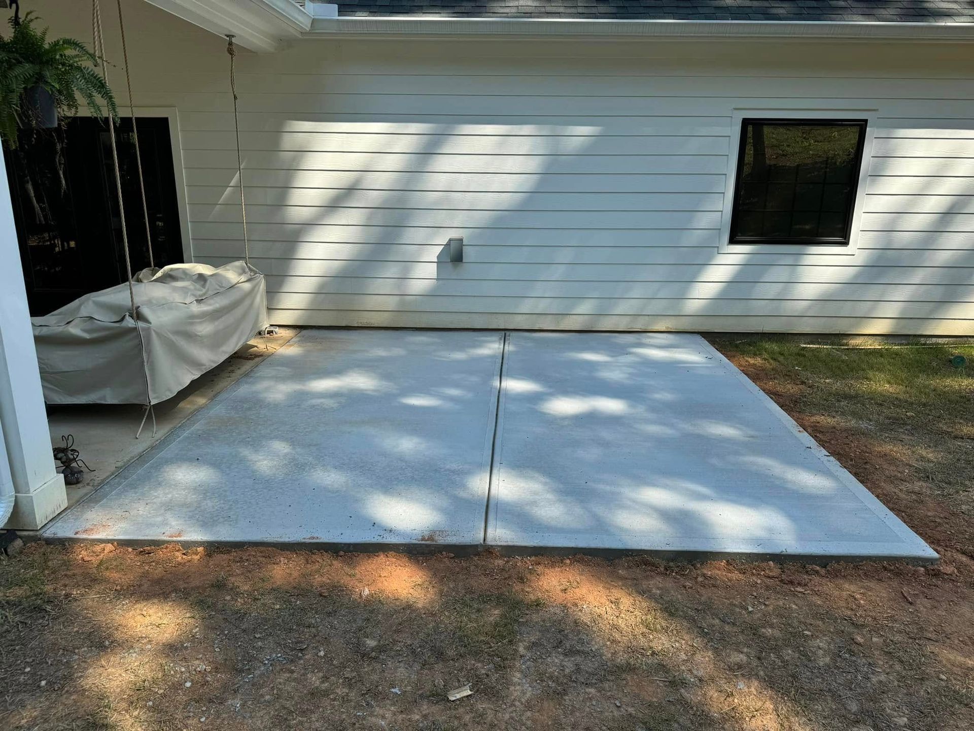 Concrete patio adjacent to a white house with a covered swing and small window.