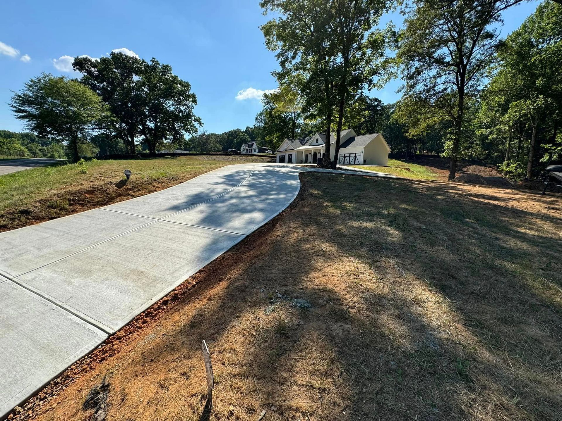 Concrete driveway leading to a light-colored house surrounded by trees under a blue sky.