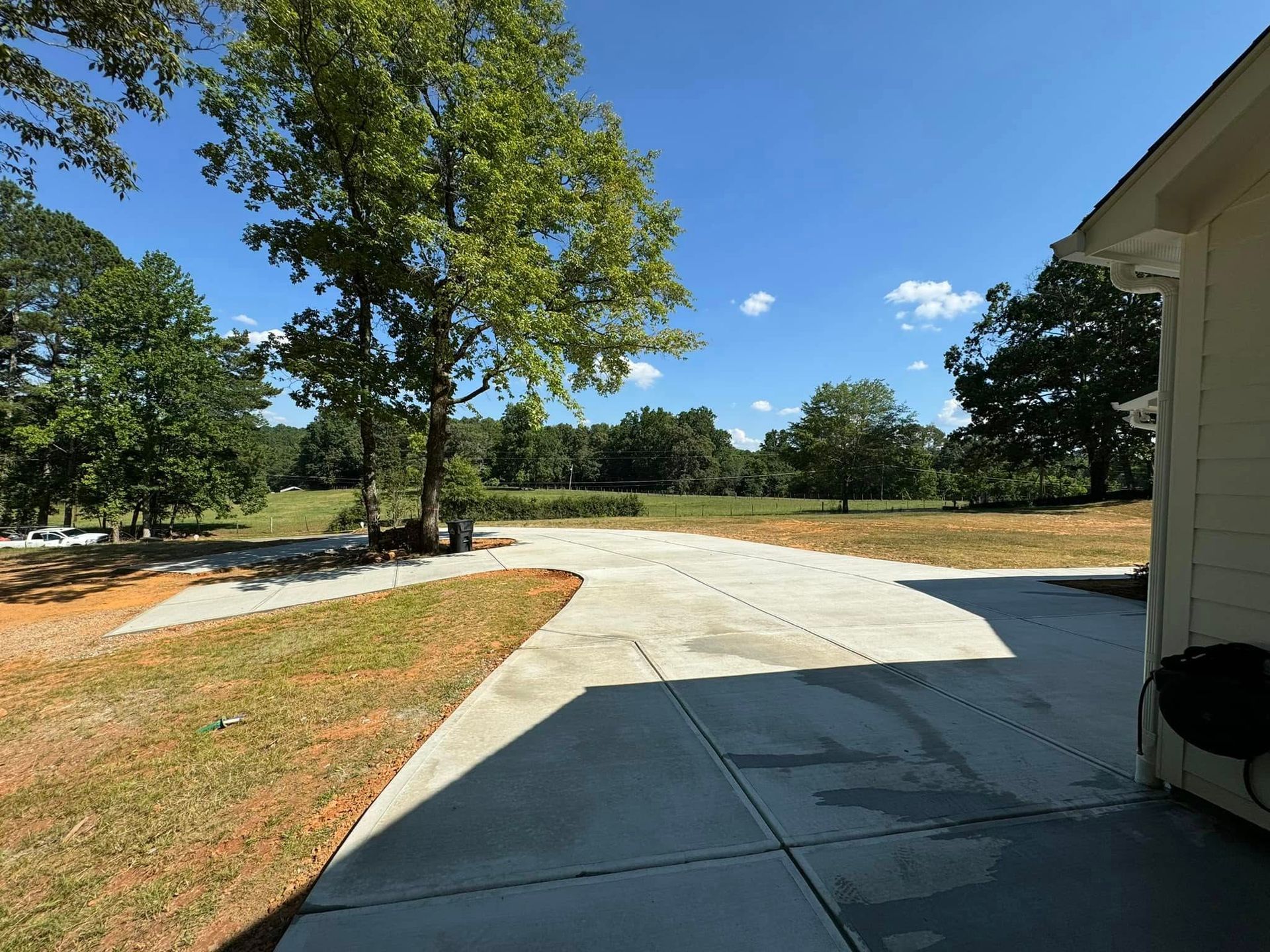 Concrete driveway leading to a grassy field under a bright blue sky with trees.