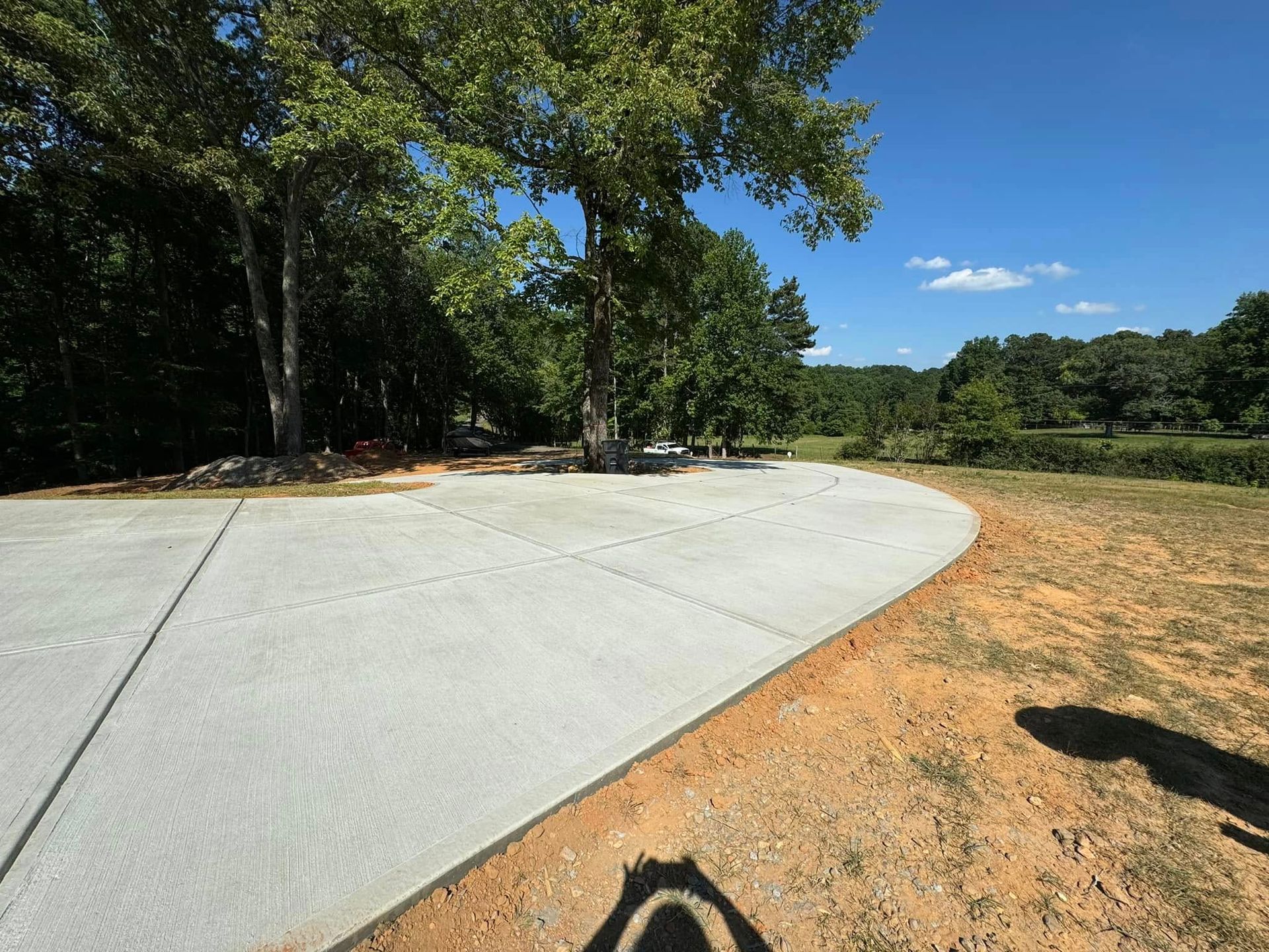A newly poured concrete driveway curves toward a tree and grassy area under a blue sky.