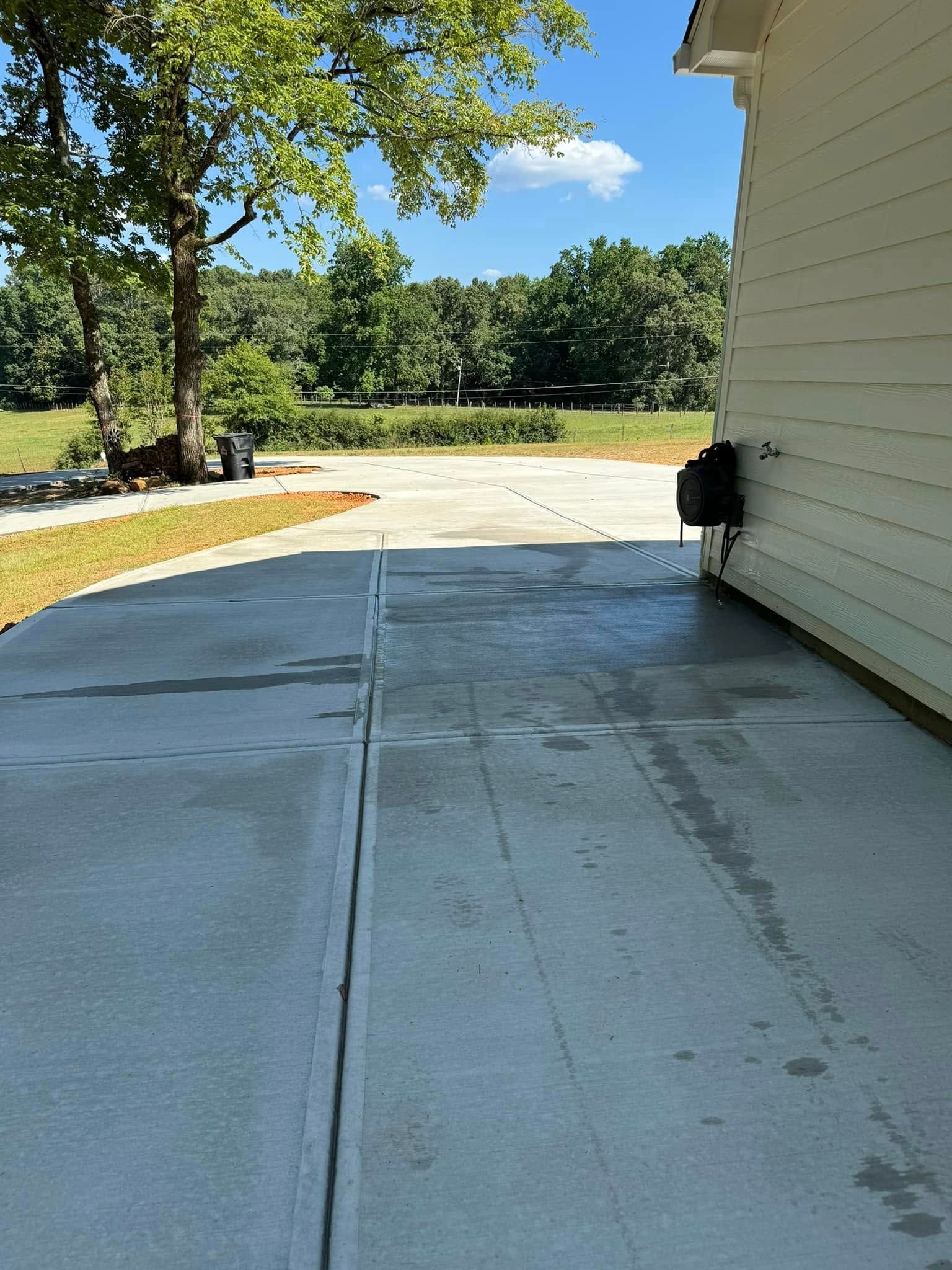 Concrete driveway next to a light-colored house. A tree is on the left; green trees and blue sky are in the distance.