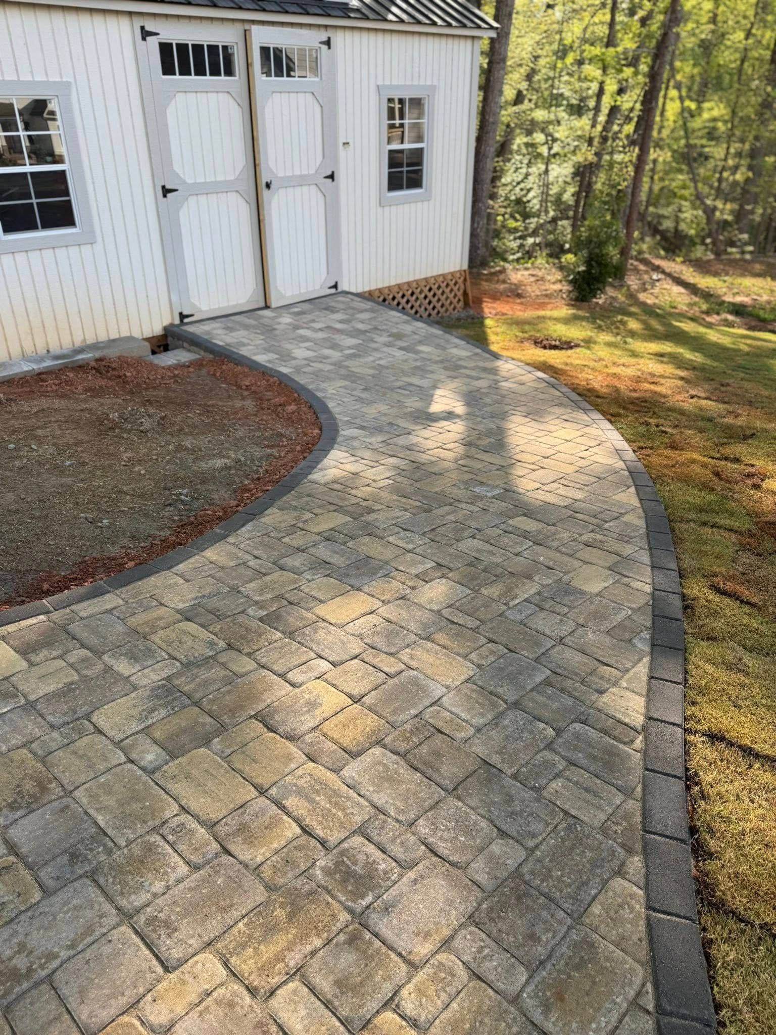 Brick pathway curves past a white shed with black accents, leading to a grassy yard.