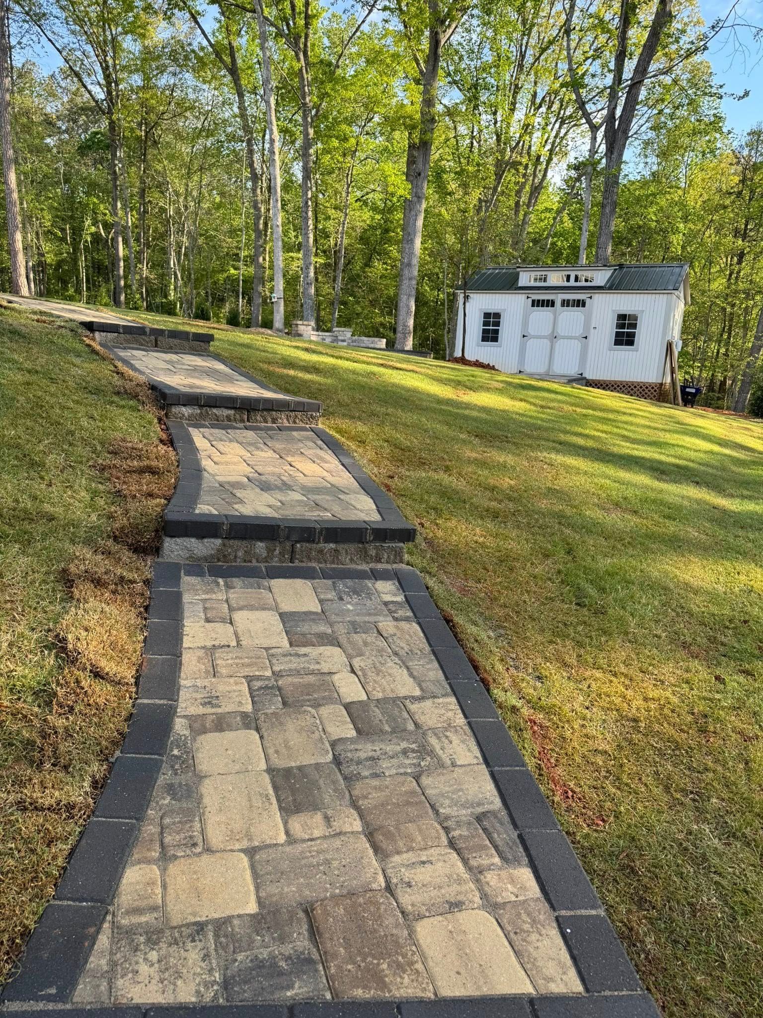Brick pathway with steps leads uphill to a shed on a grassy lawn, trees in the background.