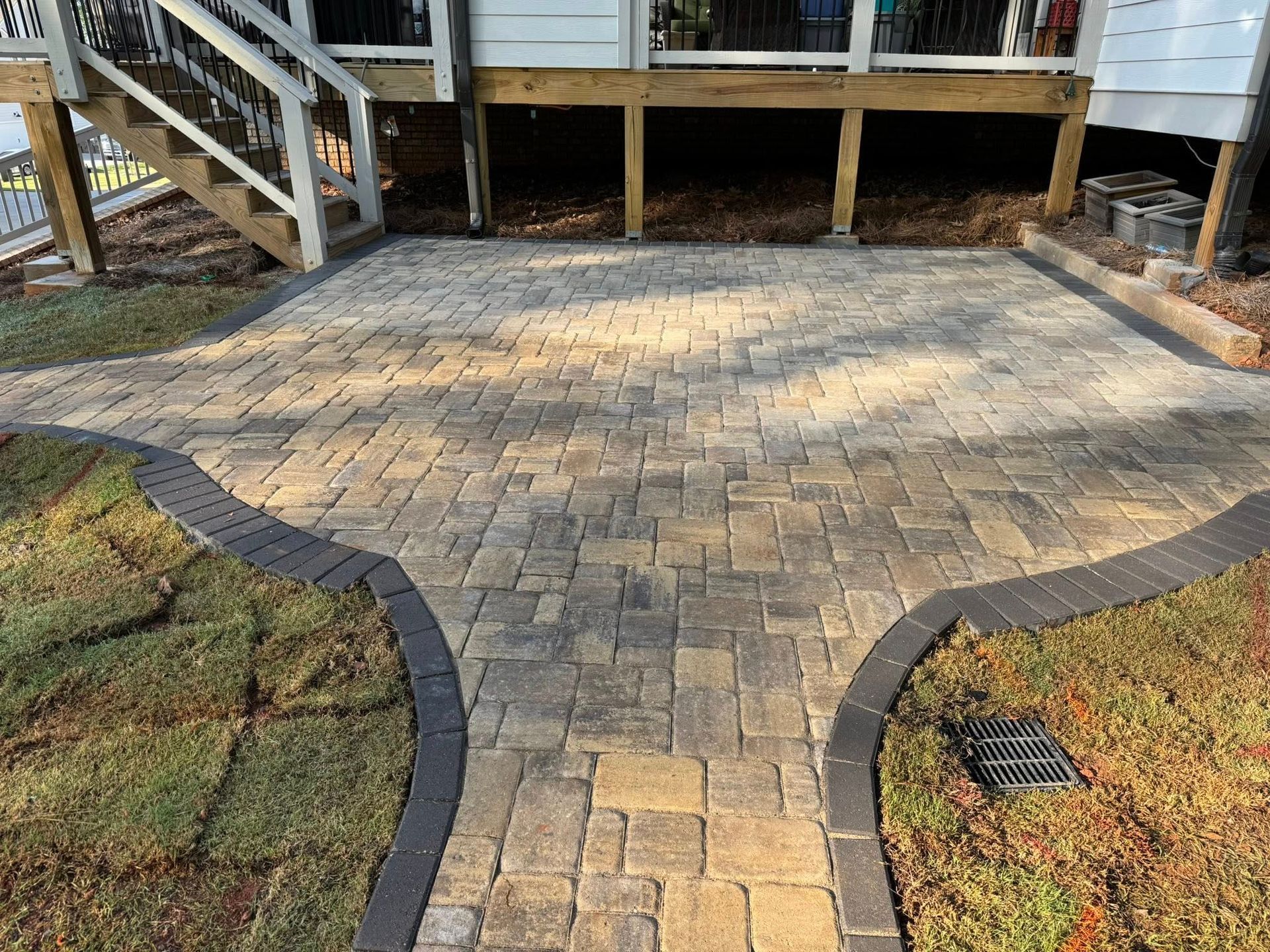 Brick patio with dark border and grass path leading to steps under a wooden deck.