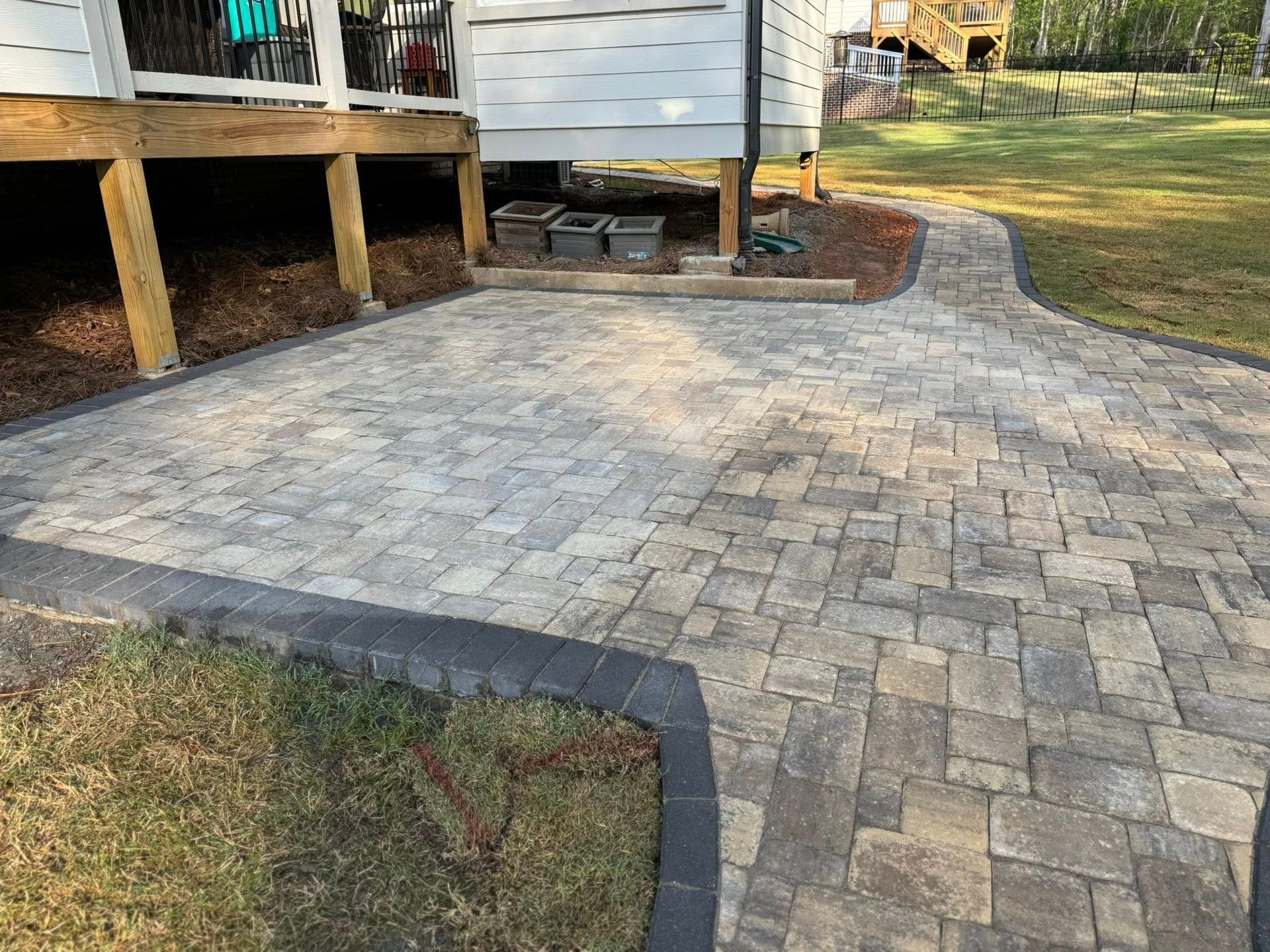 Paver patio and walkway adjacent to a deck, with dark edging and green grass.