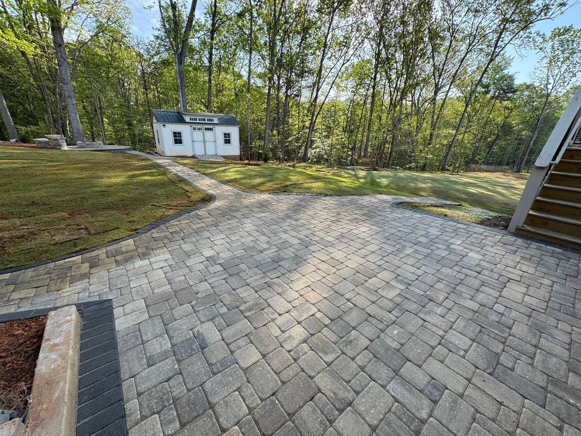 Brick patio leads to a shed in a grassy yard, surrounded by trees.