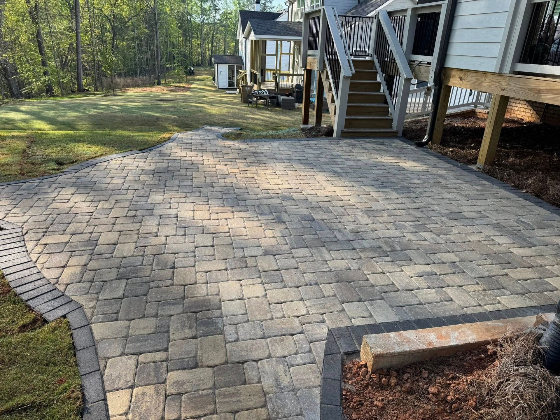 Brick patio next to a house with a wooden staircase and a small building in the background.
