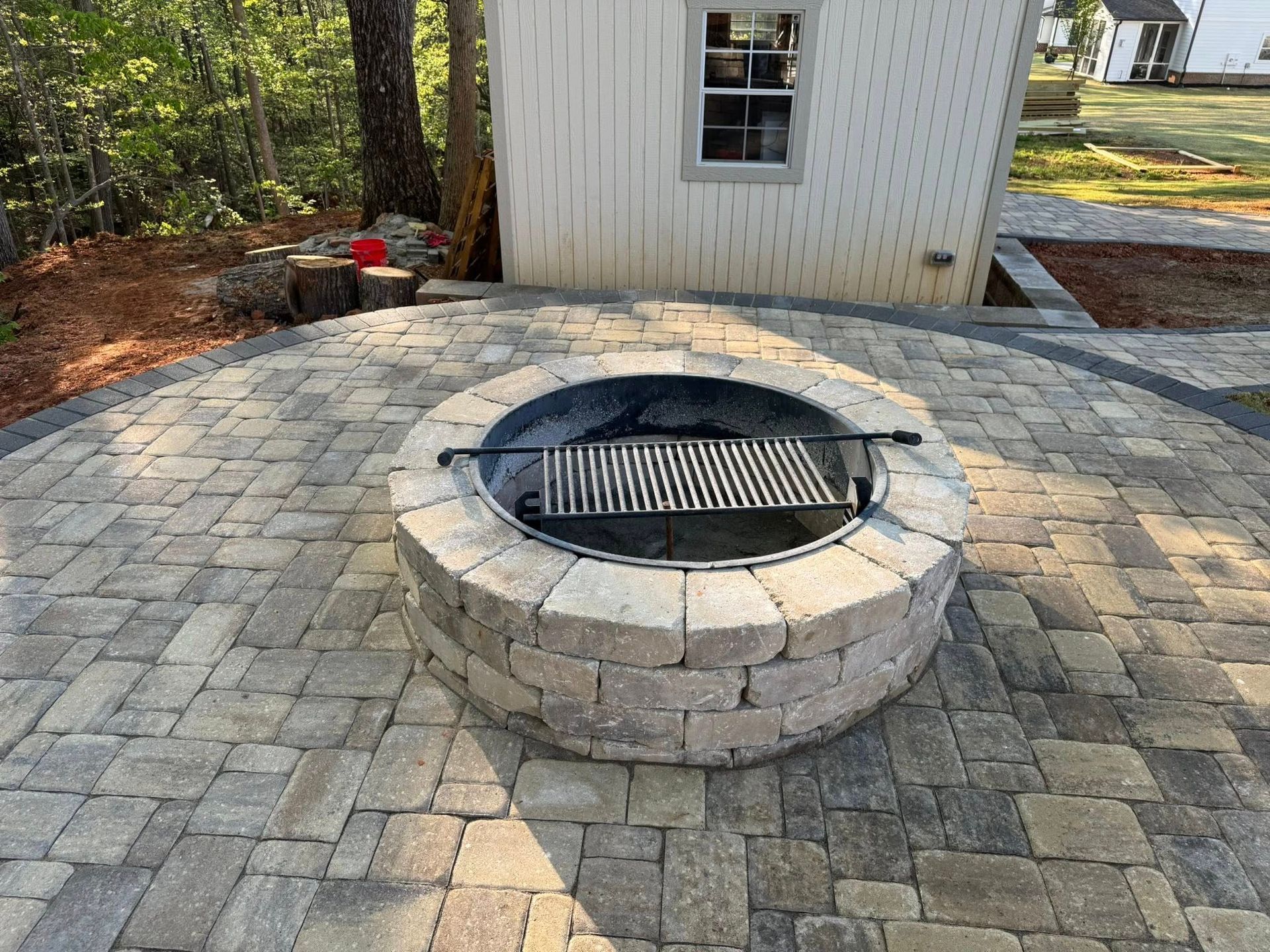 Circular stone fire pit with grill, on a brick patio, near a white shed.