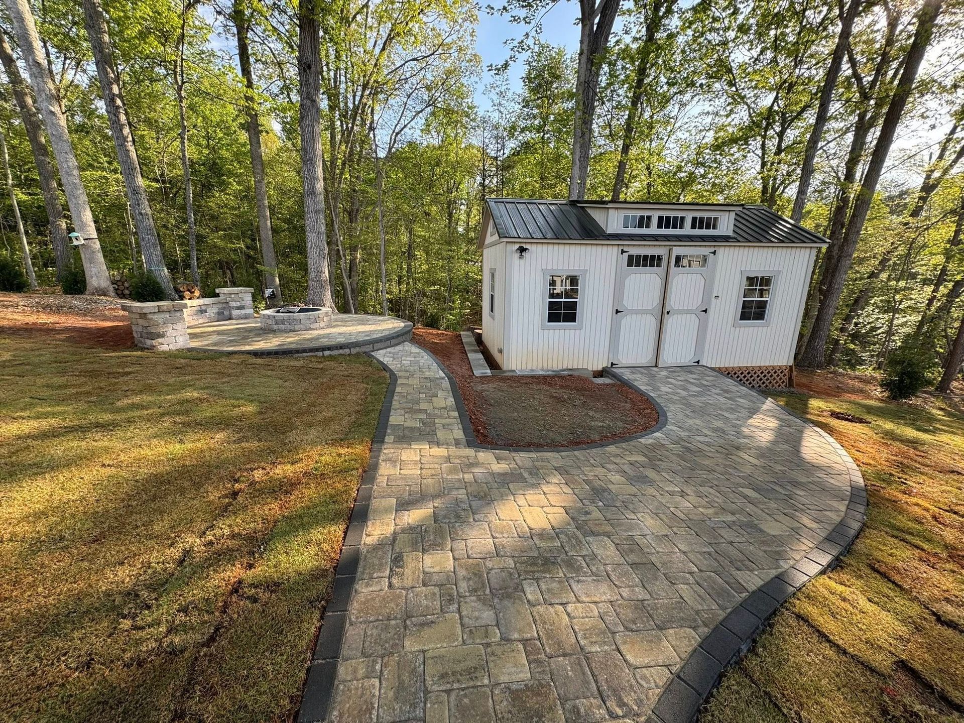 A white shed with double doors and a brick pathway surrounded by grass and trees.